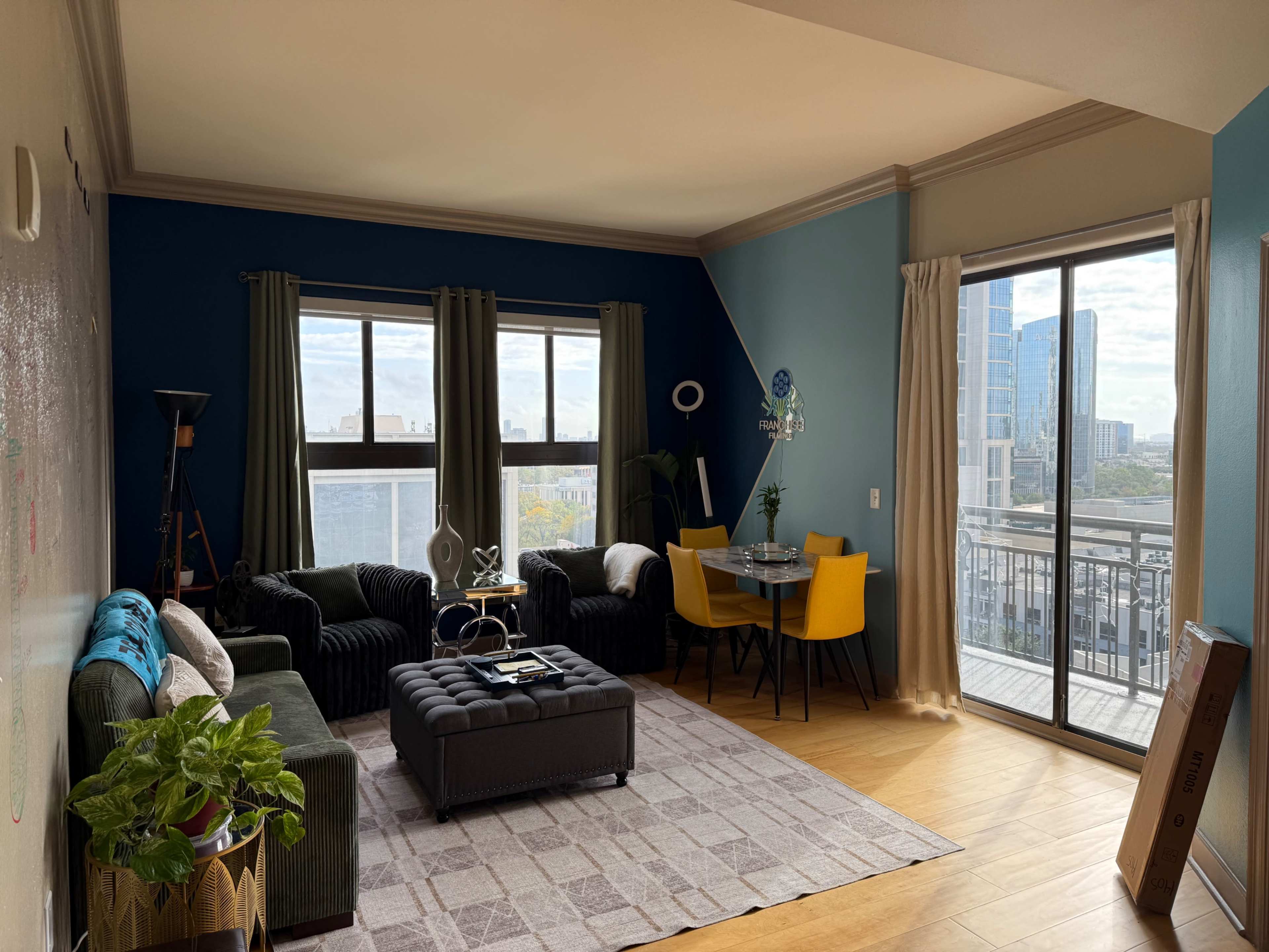 A furnished living room with a dark blue accent wall, large windows, and a balcony overlooks a city skyline.