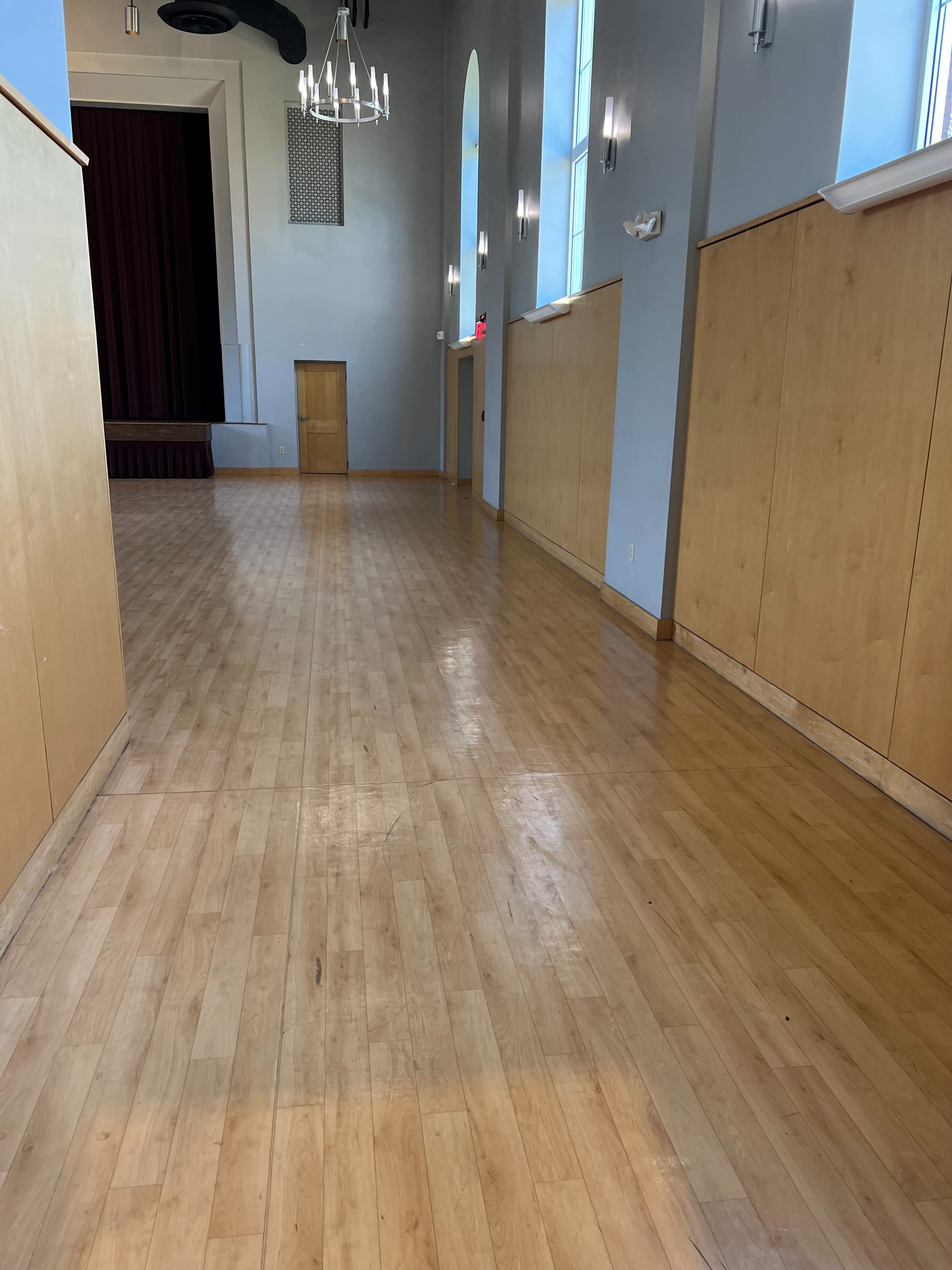 A long, wooden-floored hallway in a spacious indoor venue with large windows and a stage area at one end.