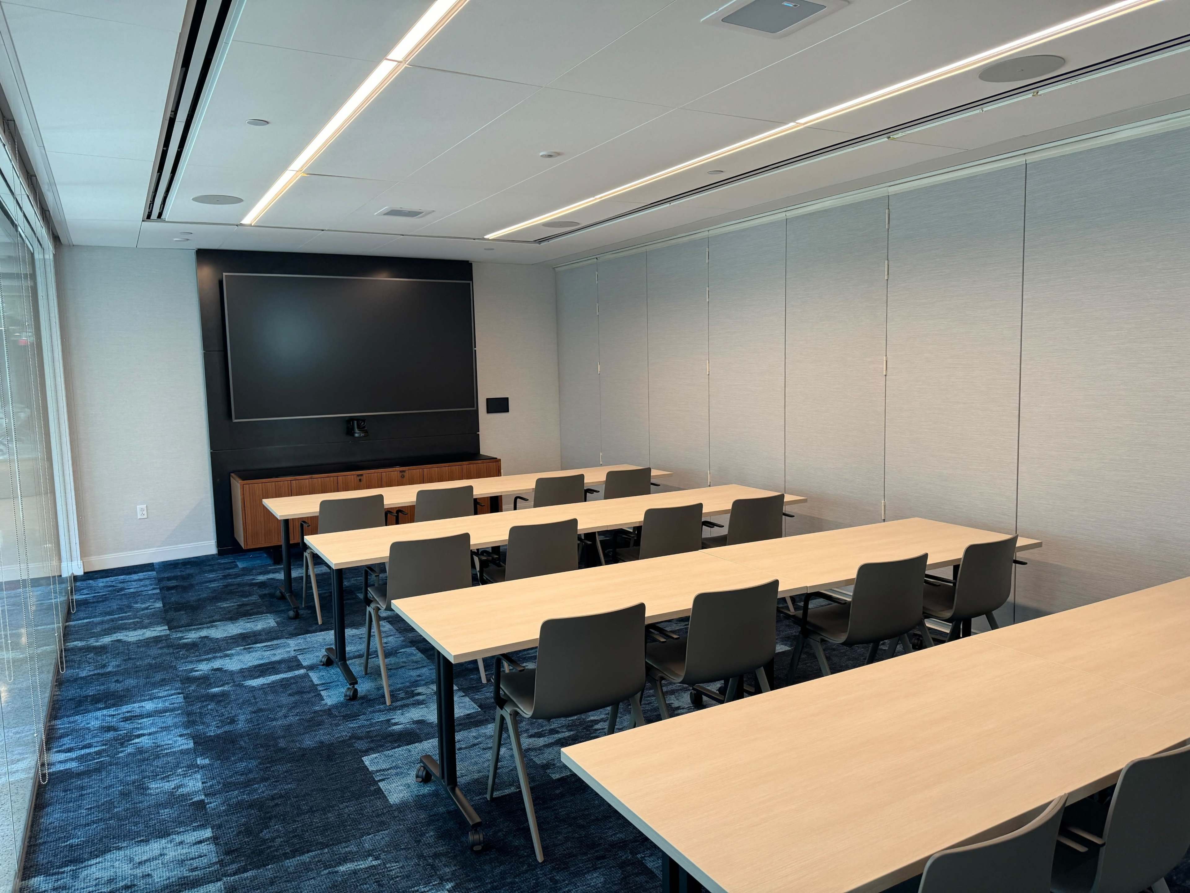 A modern conference room features multiple long tables arranged with chairs facing a wall-mounted screen.