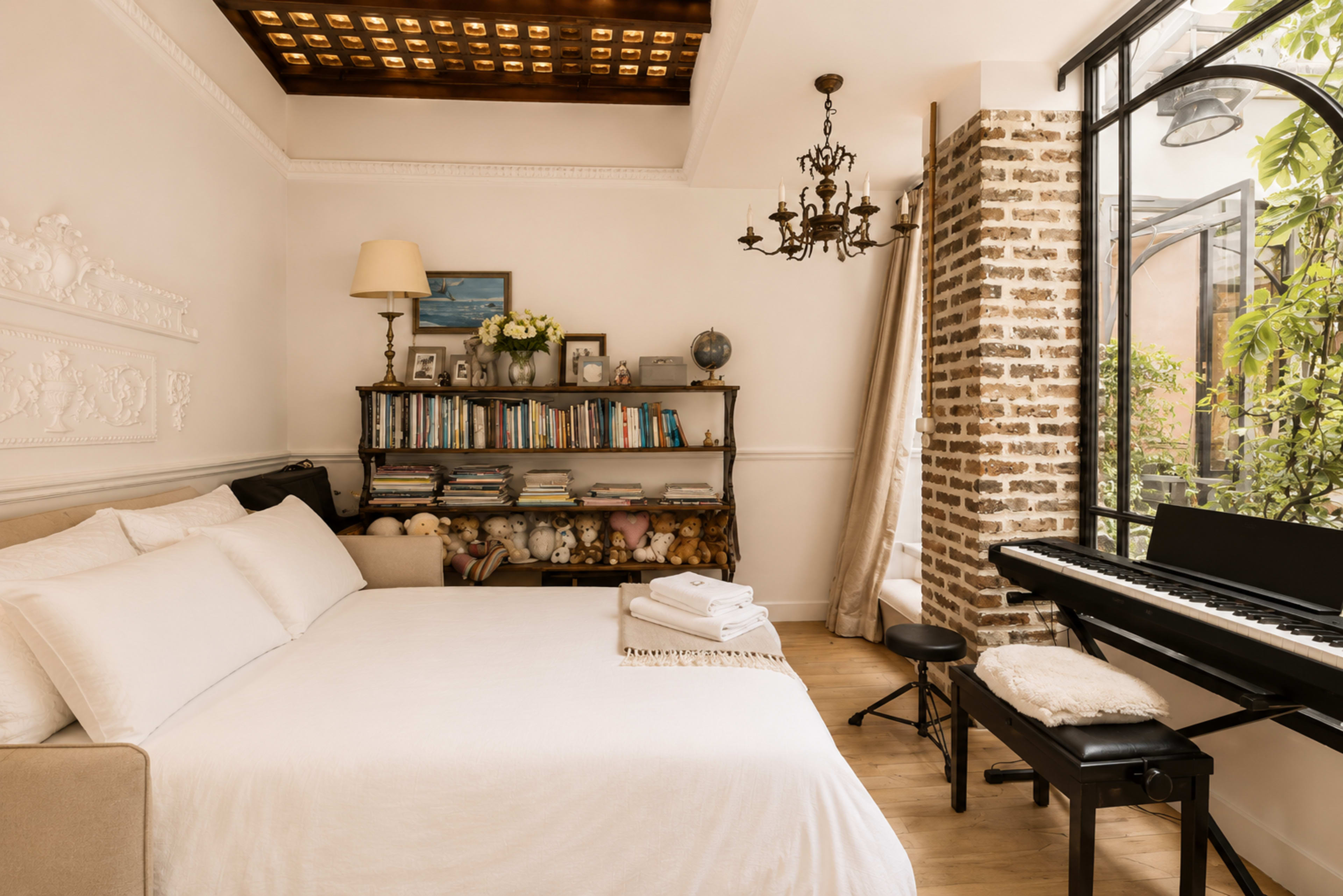 The image shows a cozy bedroom with a bed, bookshelves filled with books and stuffed animals, a piano, and large windows that allow natural light to enter.