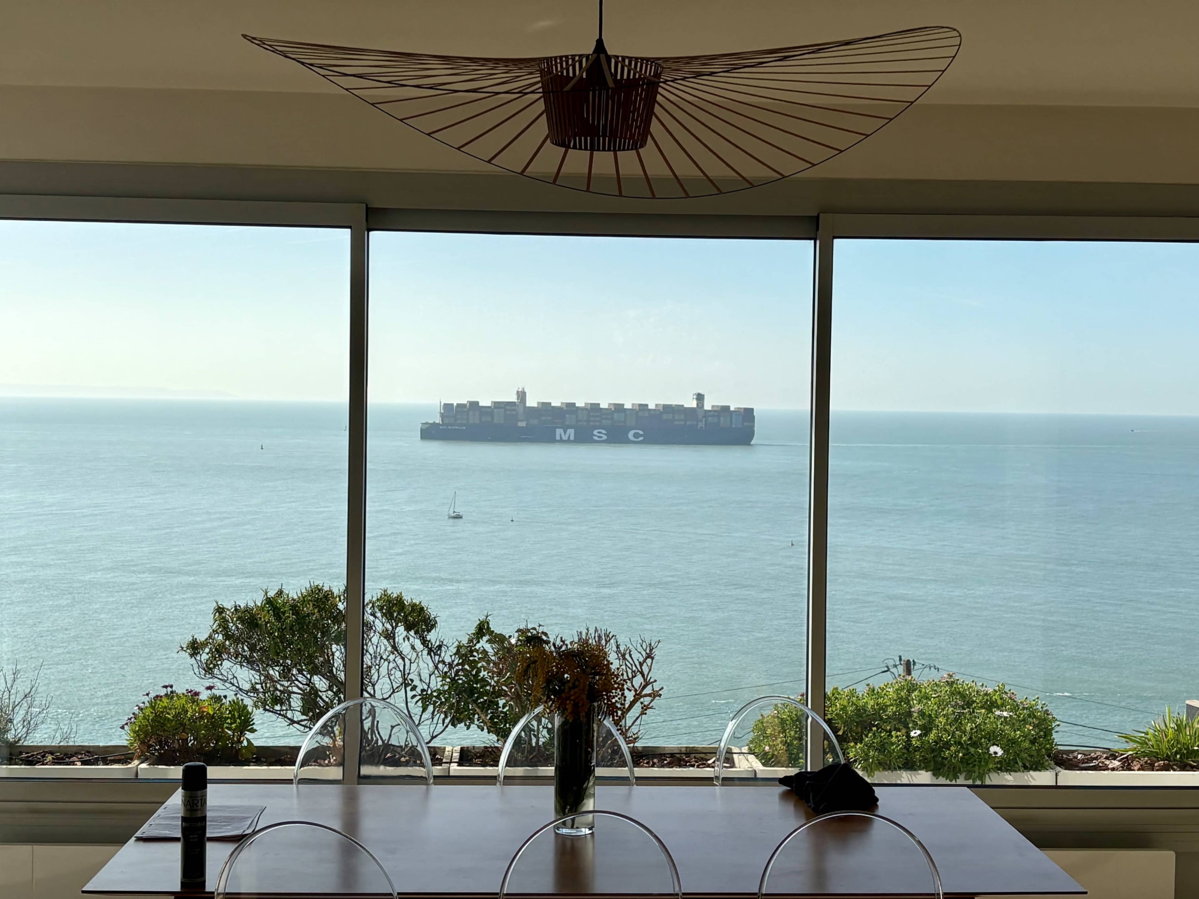 A large container ship is anchored in the water, visible through a large window with a dining table and plants in the foreground.