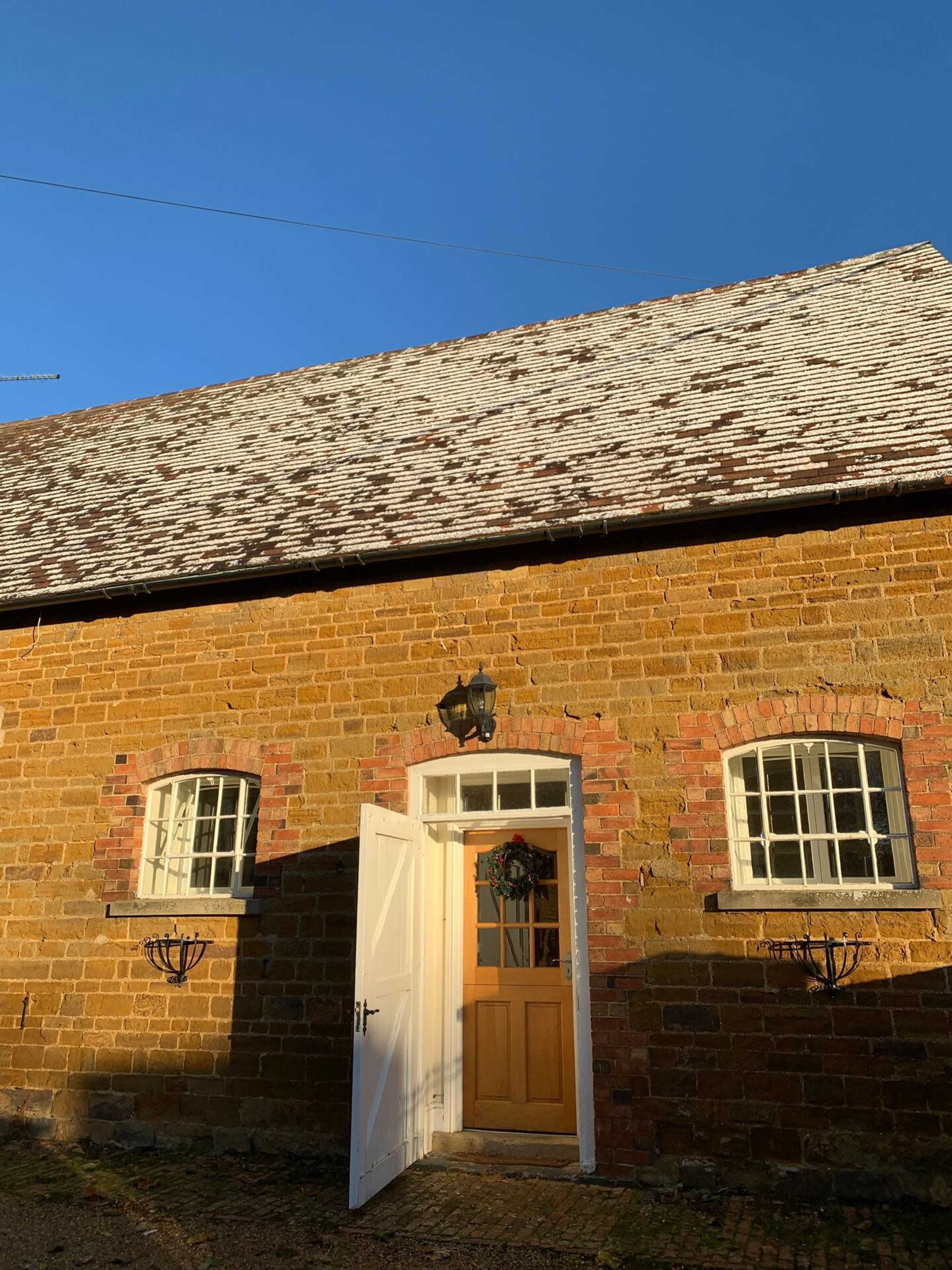 A stone cottage with a wooden door and two windows, featuring a sloped roof, stands against a clear blue sky.