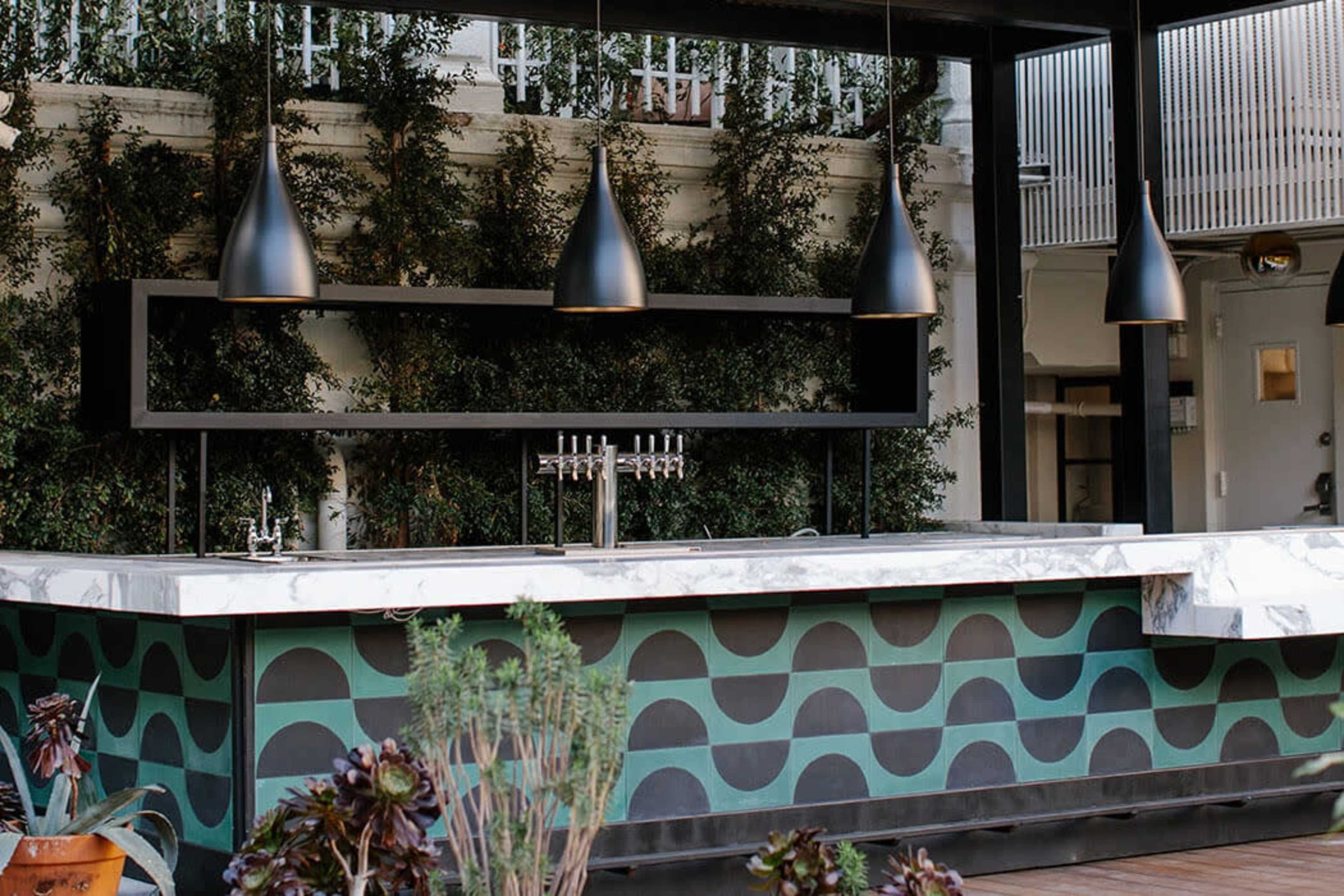 The image shows an outdoor bar with a patterned counter, black pendant lights hanging above, and greenery in the background.