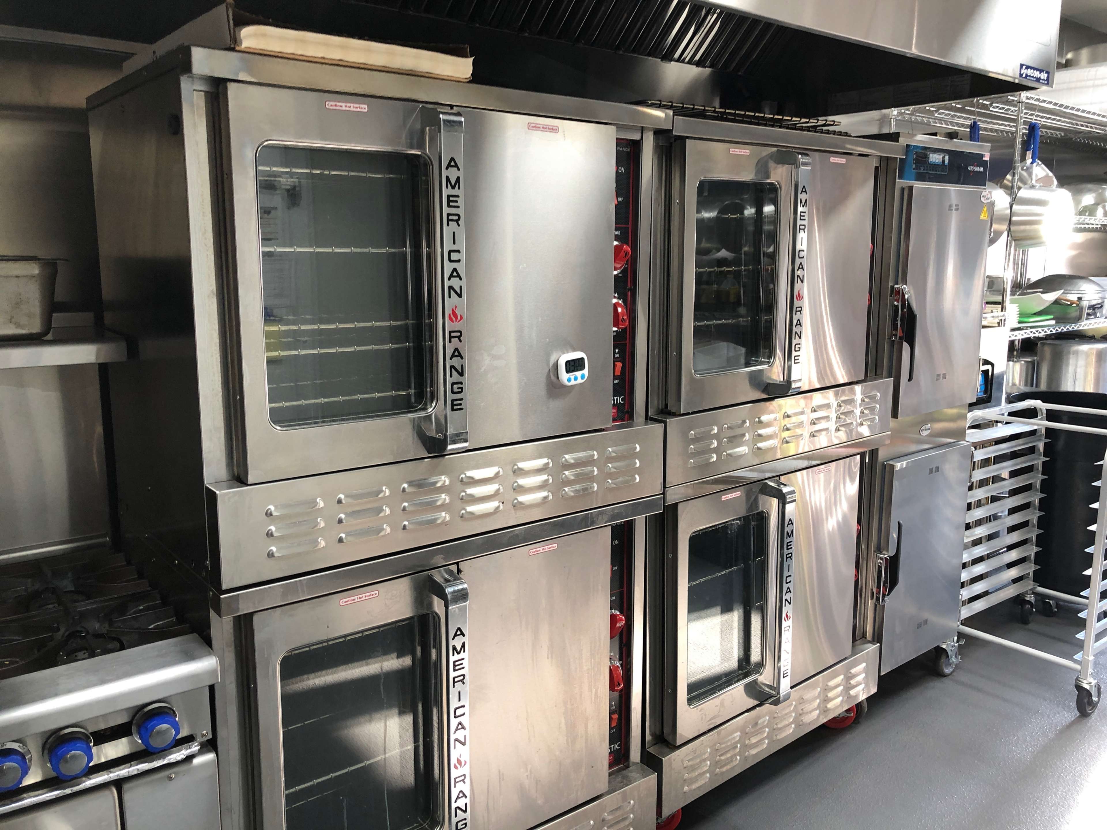 The image shows a commercial kitchen setup featuring a stacked unit of stainless steel convection ovens with a separate refrigerator on the side.