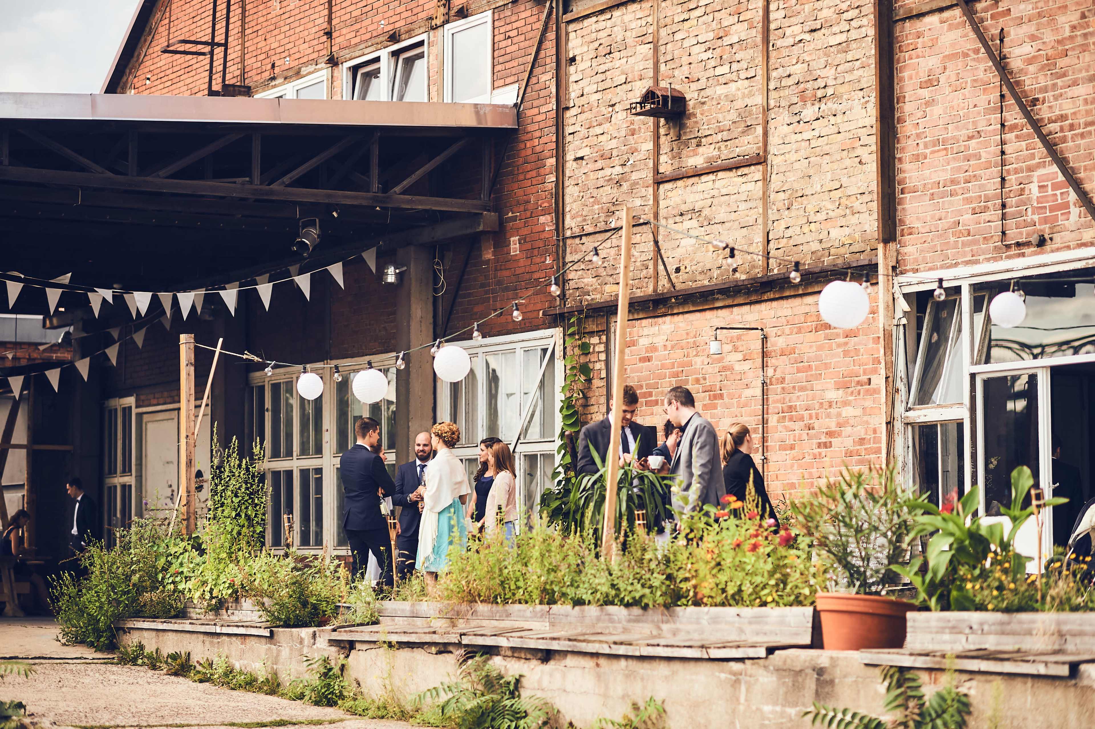 A group of people socializes outside a rustic brick building adorned with plants and string lights.