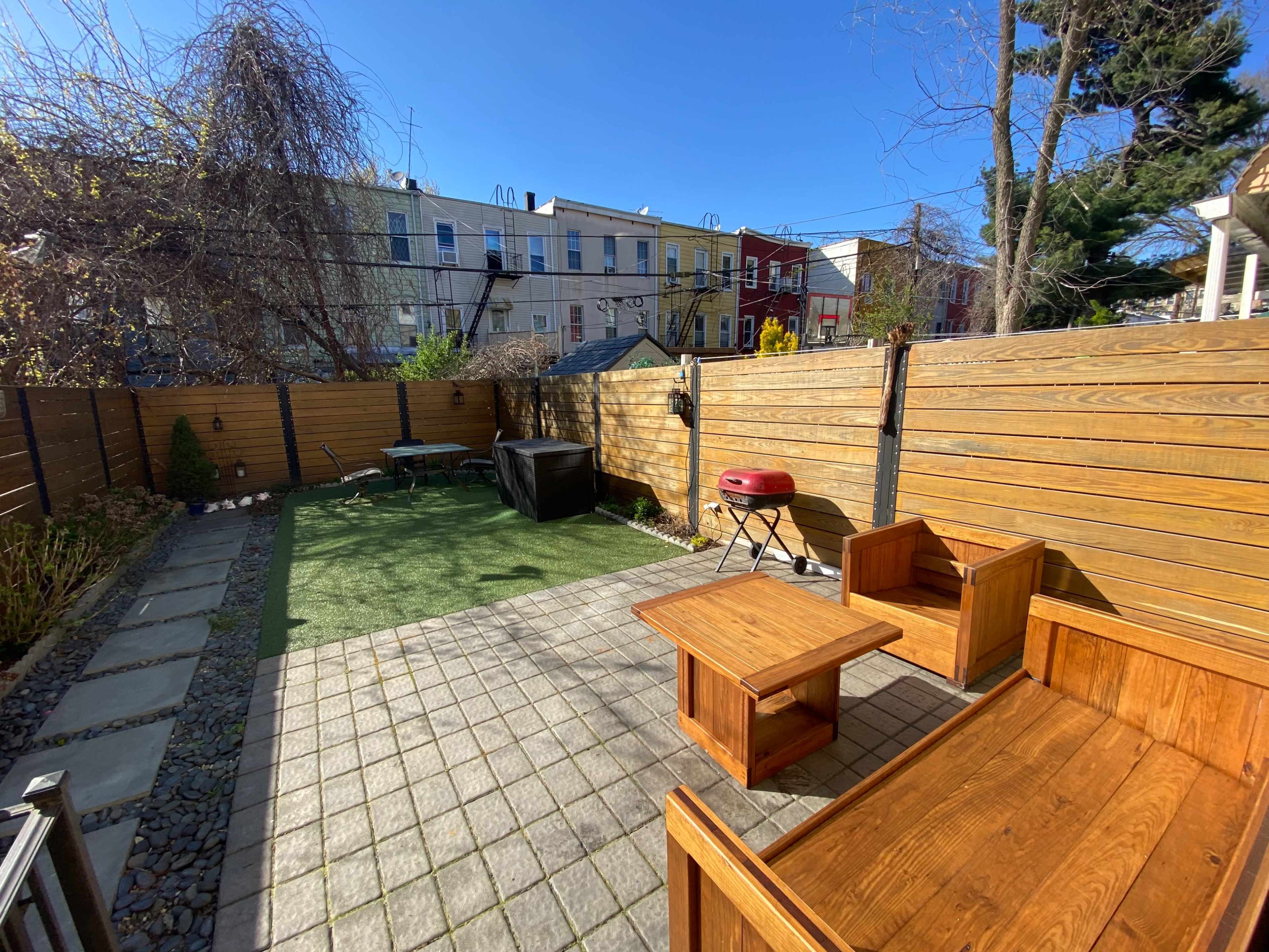 The outdoor patio features wooden furniture, a barbecue grill, and a small artificial grass area, surrounded by wooden fencing and neighboring buildings.