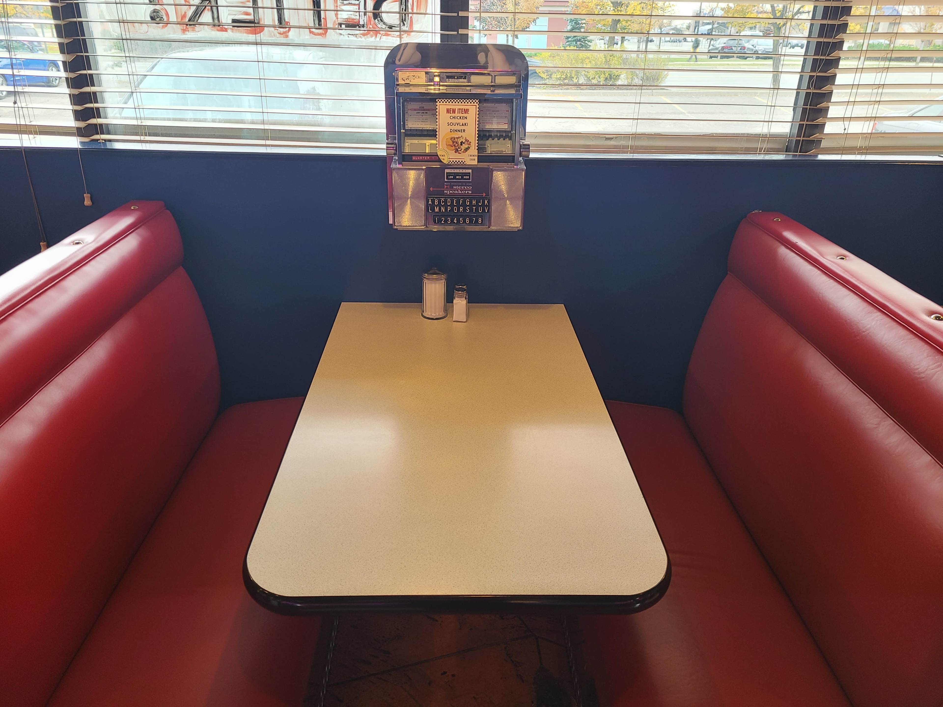 A red vinyl booth with a white tabletop is set up in a diner, featuring a vintage jukebox and condiments on the table.