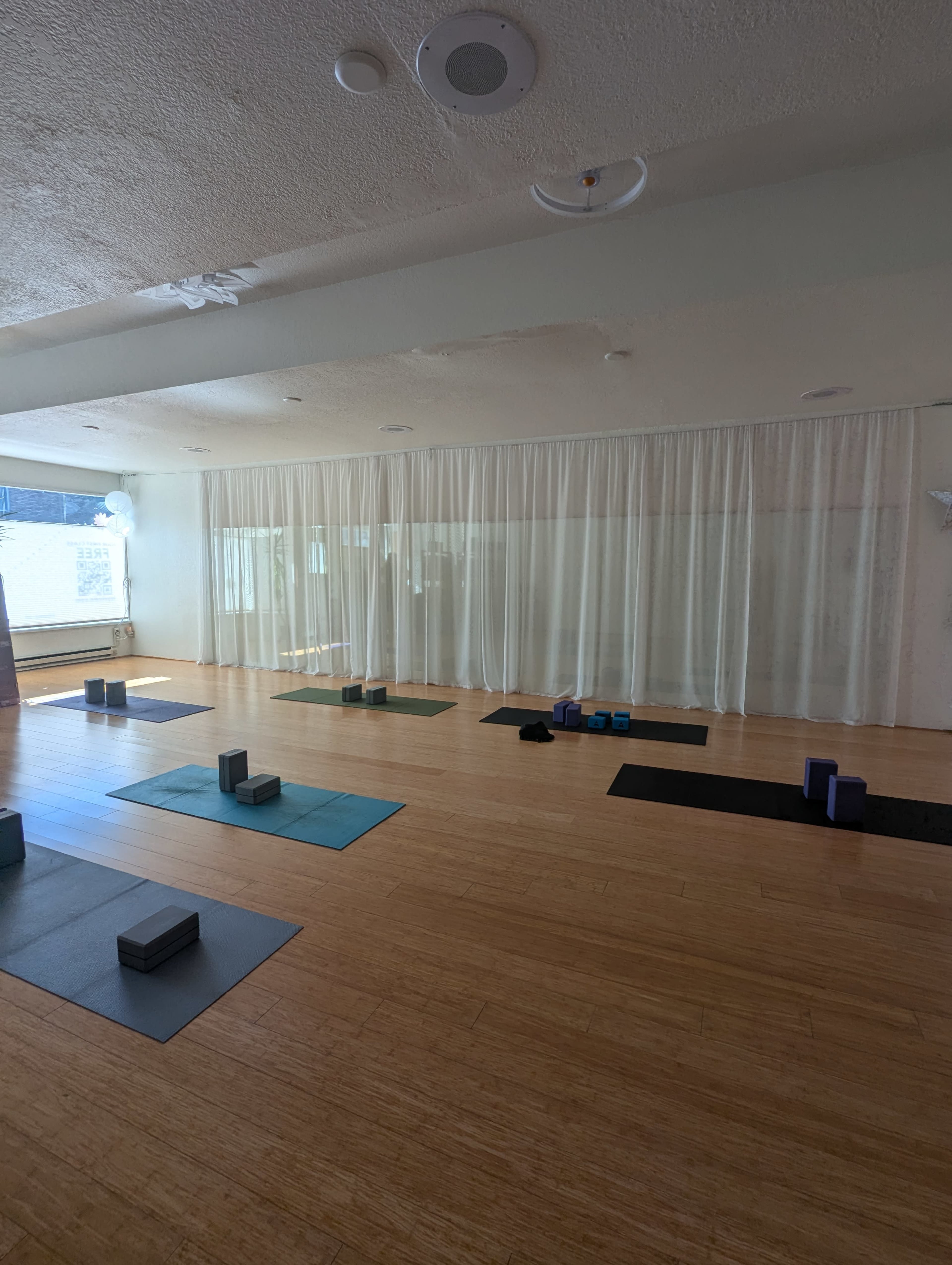 A spacious yoga studio features bamboo flooring, exercise mats, and props arranged neatly, with sheer curtains allowing natural light to filter in.