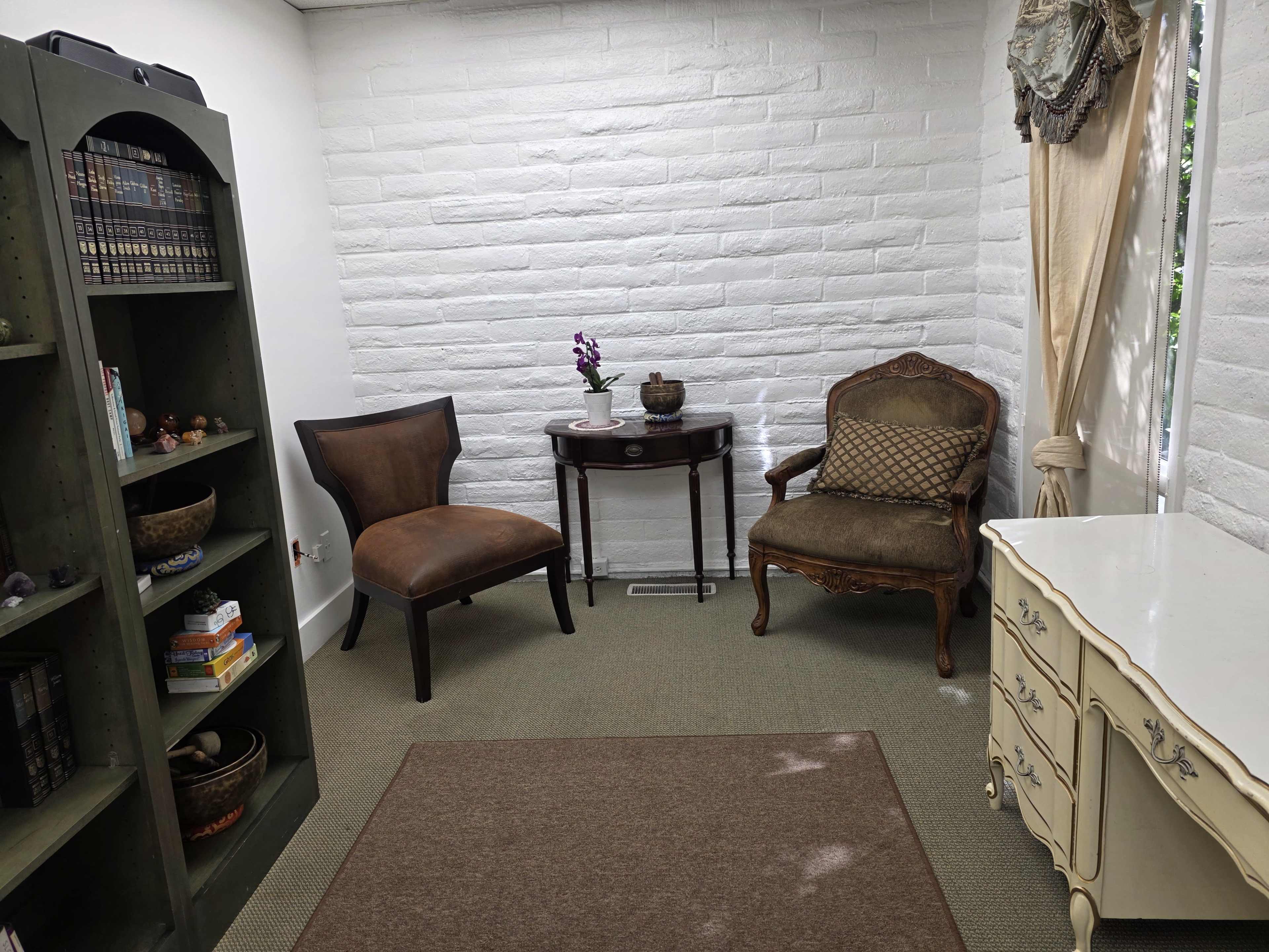 The image shows a small, neat room with a bookshelf, two chairs, a table with a decorative item, and a dresser against a white brick wall.