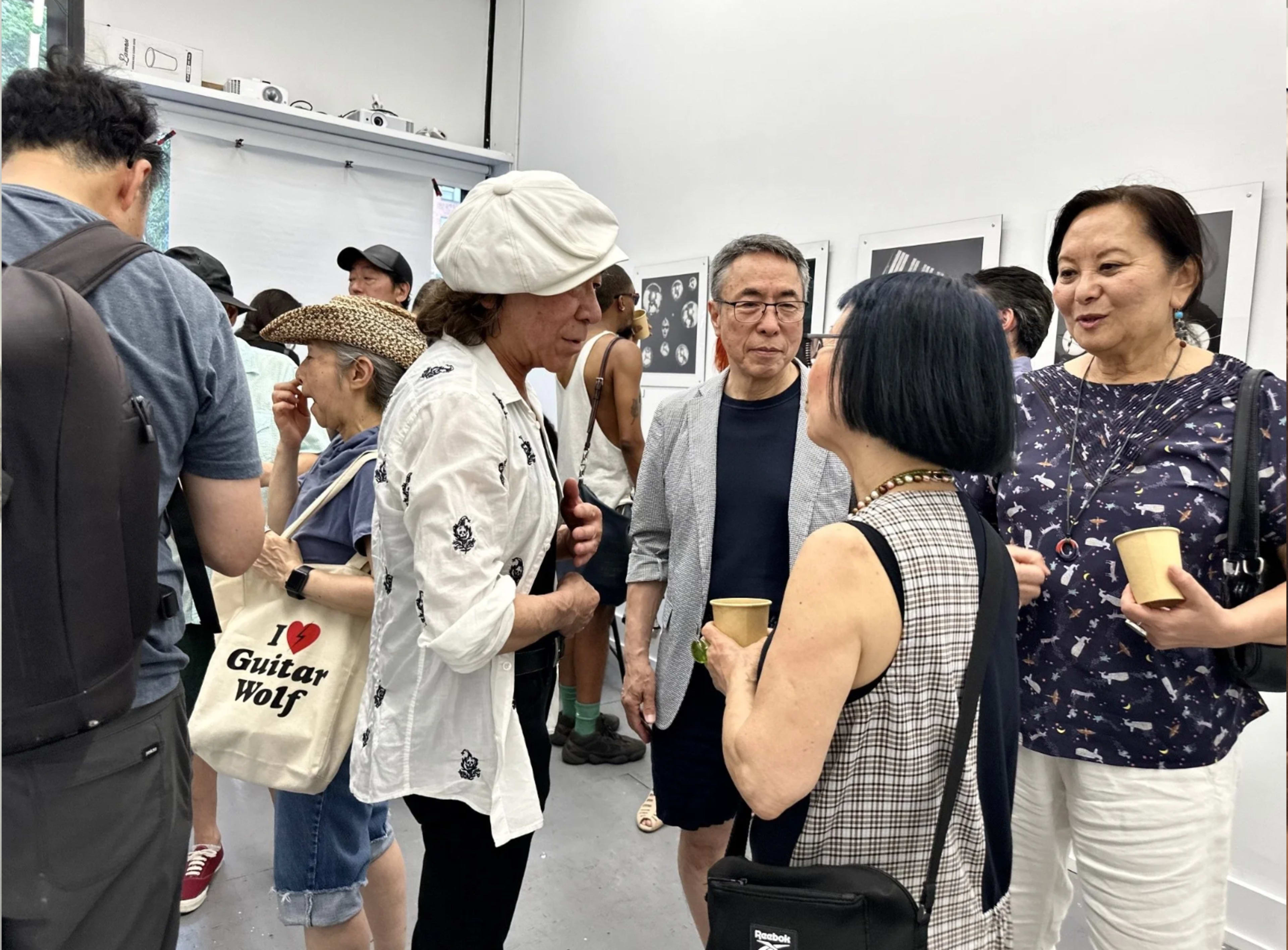 A group of people are engaged in conversation at an art event in a gallery, surrounded by black-and-white photographs on the walls.