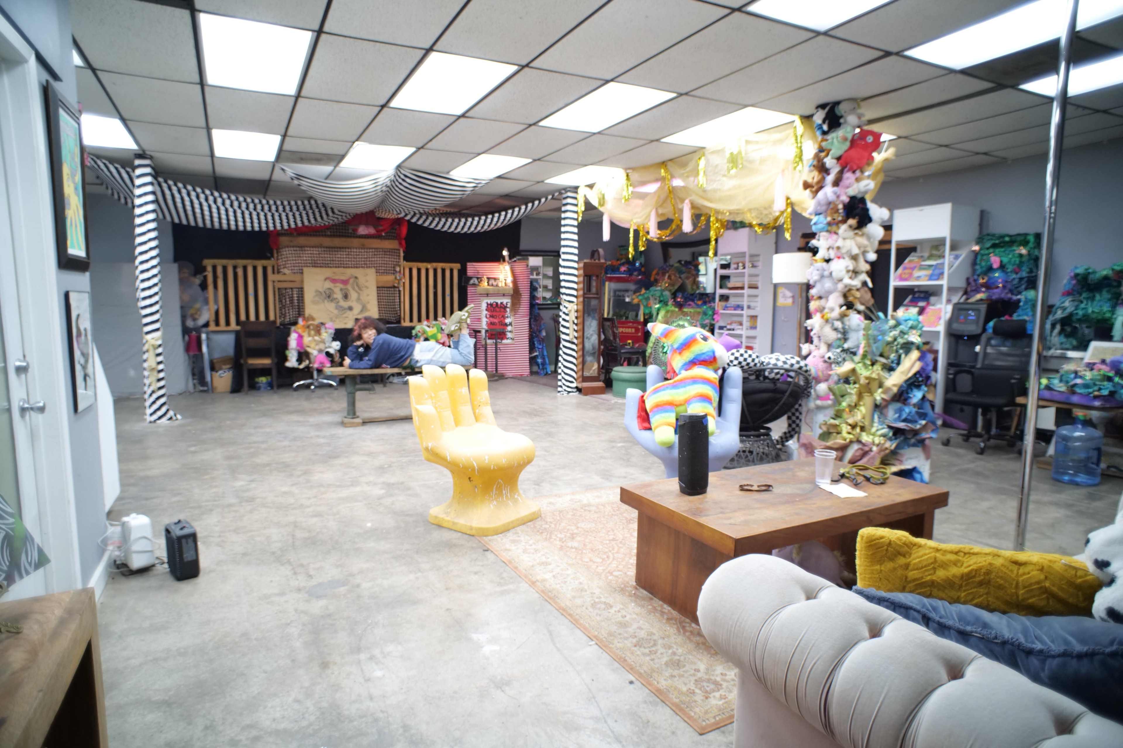 The image shows a vibrant art studio or creative space filled with colorful decorations, various seating arrangements, and art materials scattered throughout.