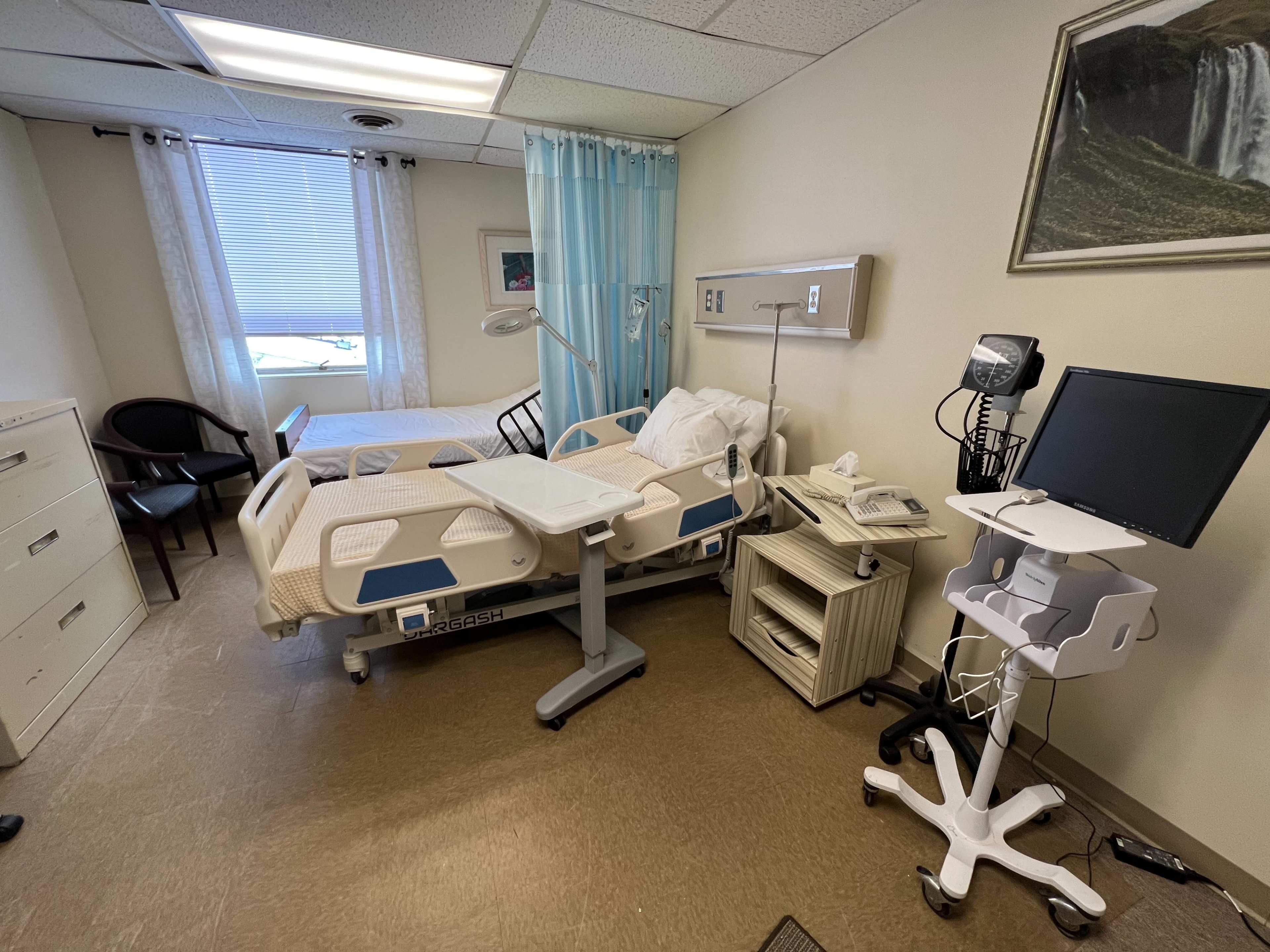 The image shows a hospital room with a adjustable bed, a small table, a computer workstation, and a window providing natural light.