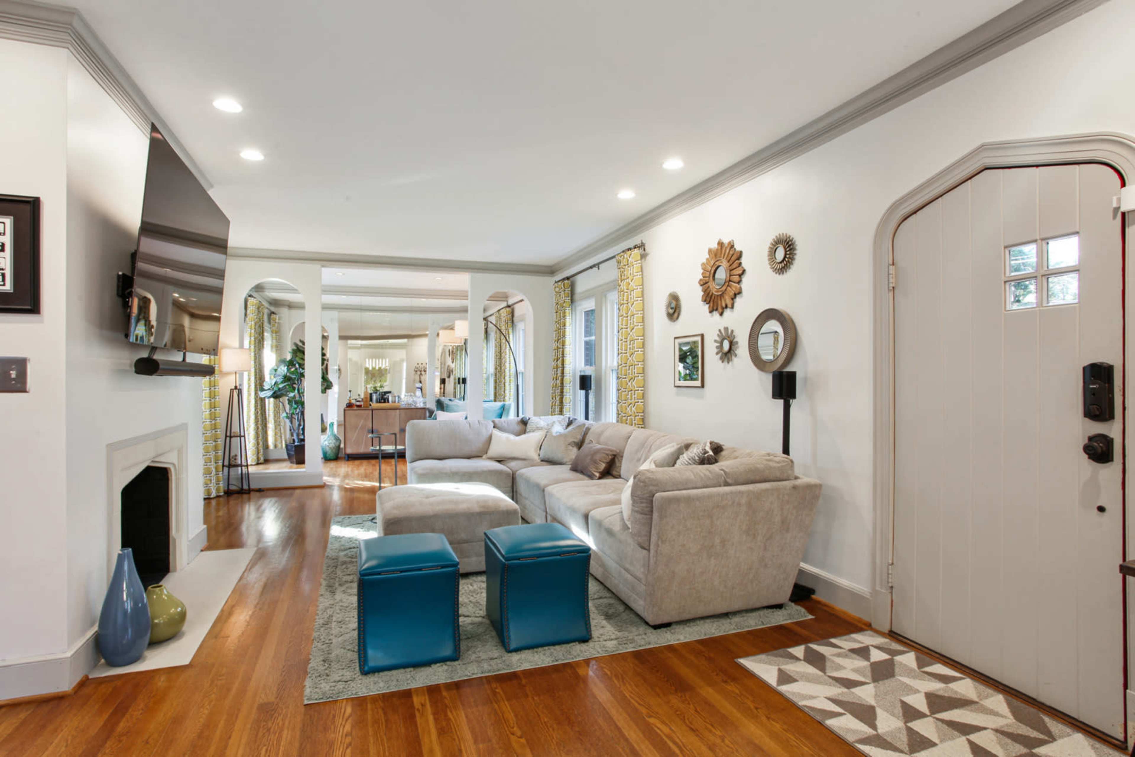 The image shows a living room with a large gray sectional sofa, a pair of blue ottomans, a television mounted on the wall, and a view into a dining area with a modern table and chairs.