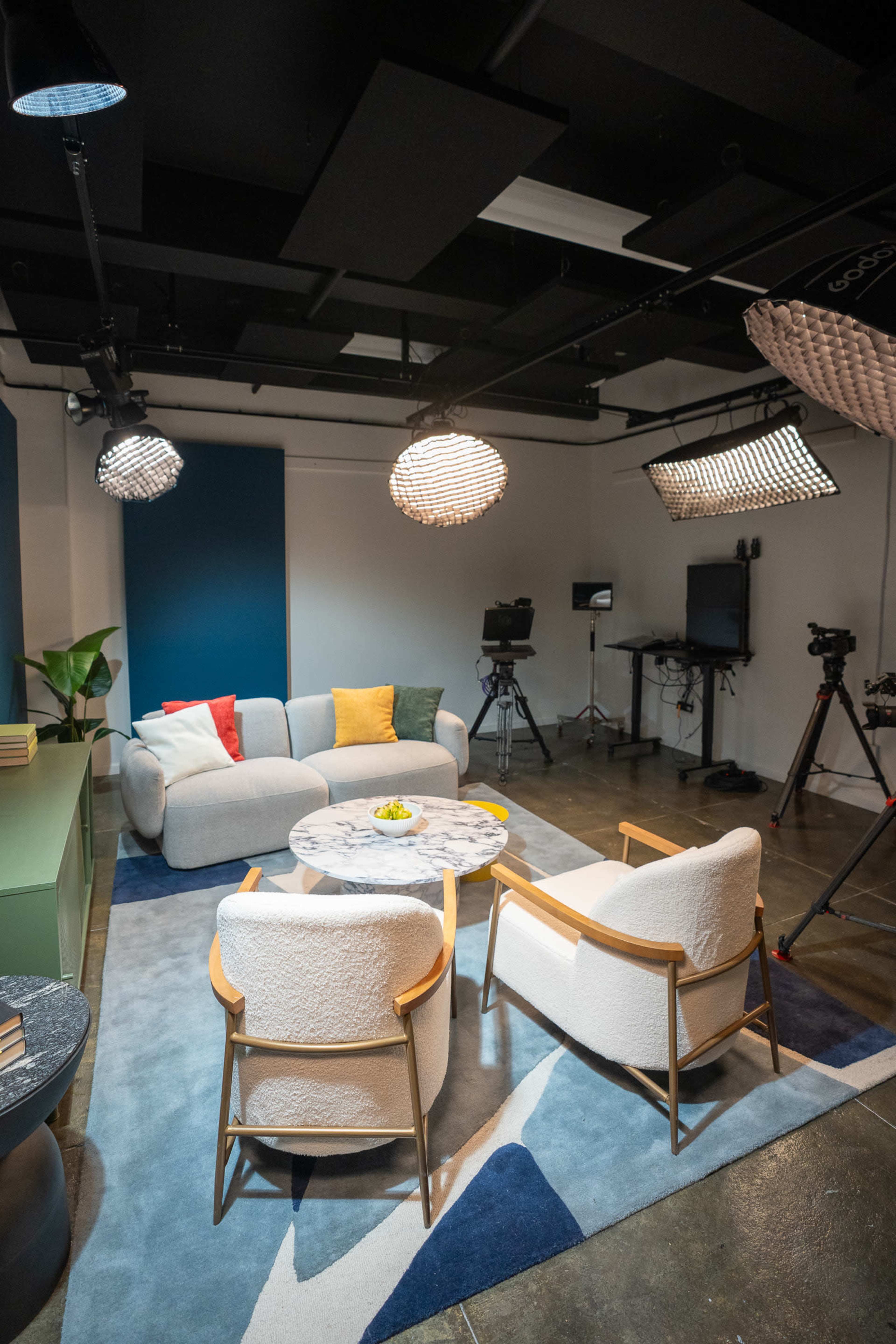 The image shows a modern studio setup featuring a circular sofa, two round chairs, a marble coffee table, and several cameras on tripods, with pendant lights hanging above and a patterned rug on the floor.