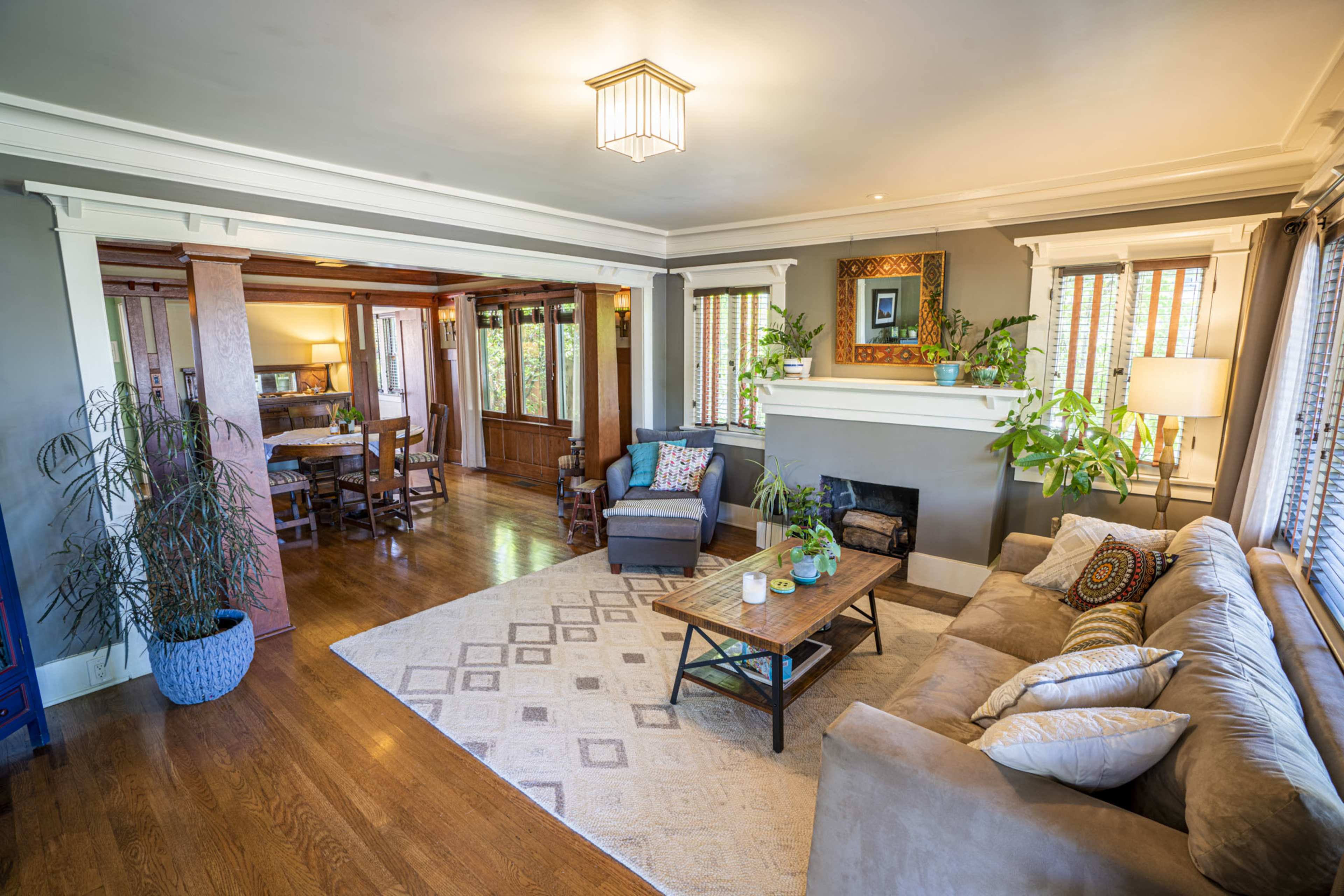 The living room features a cozy sectional sofa, a wooden coffee table, and large windows allowing natural light, with a dining area visible in the background.