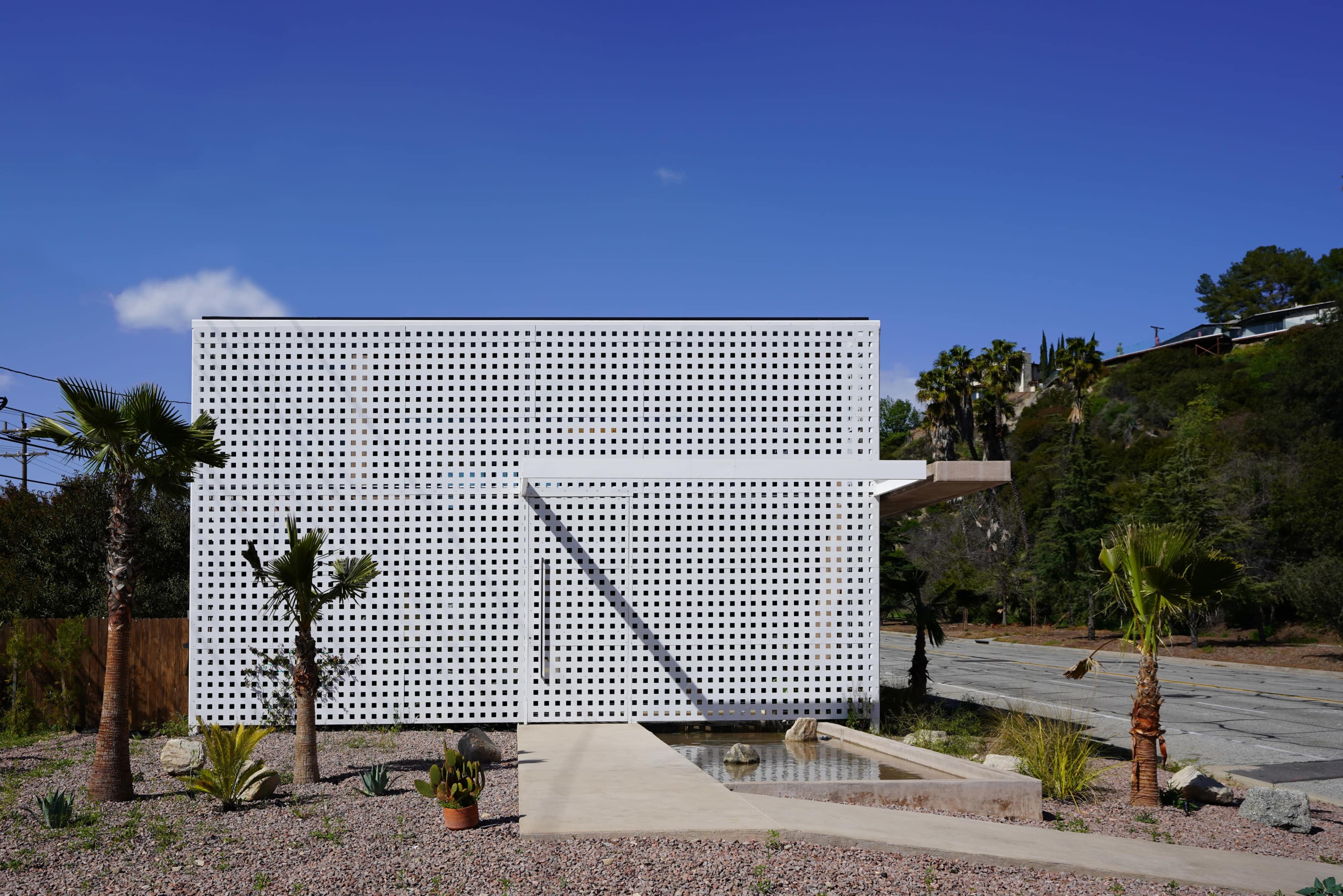 A modern, white, perforated building surrounded by a landscape of gravel and sparse greenery under a clear blue sky.