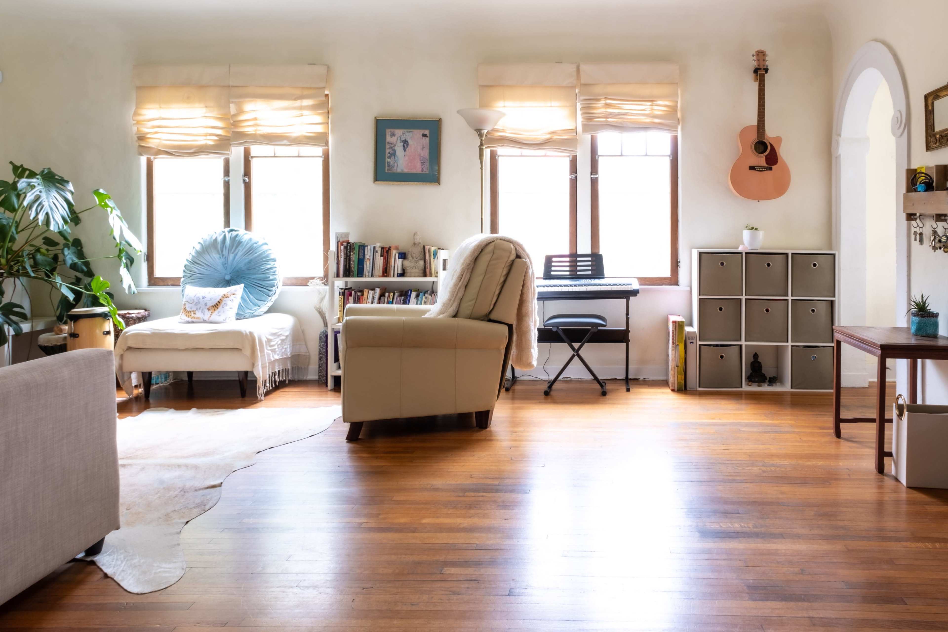 The image shows a cozy living room featuring a chair, bookshelves, a keyboard on a stand, and a guitar hanging on the wall, with natural light coming through the windows.