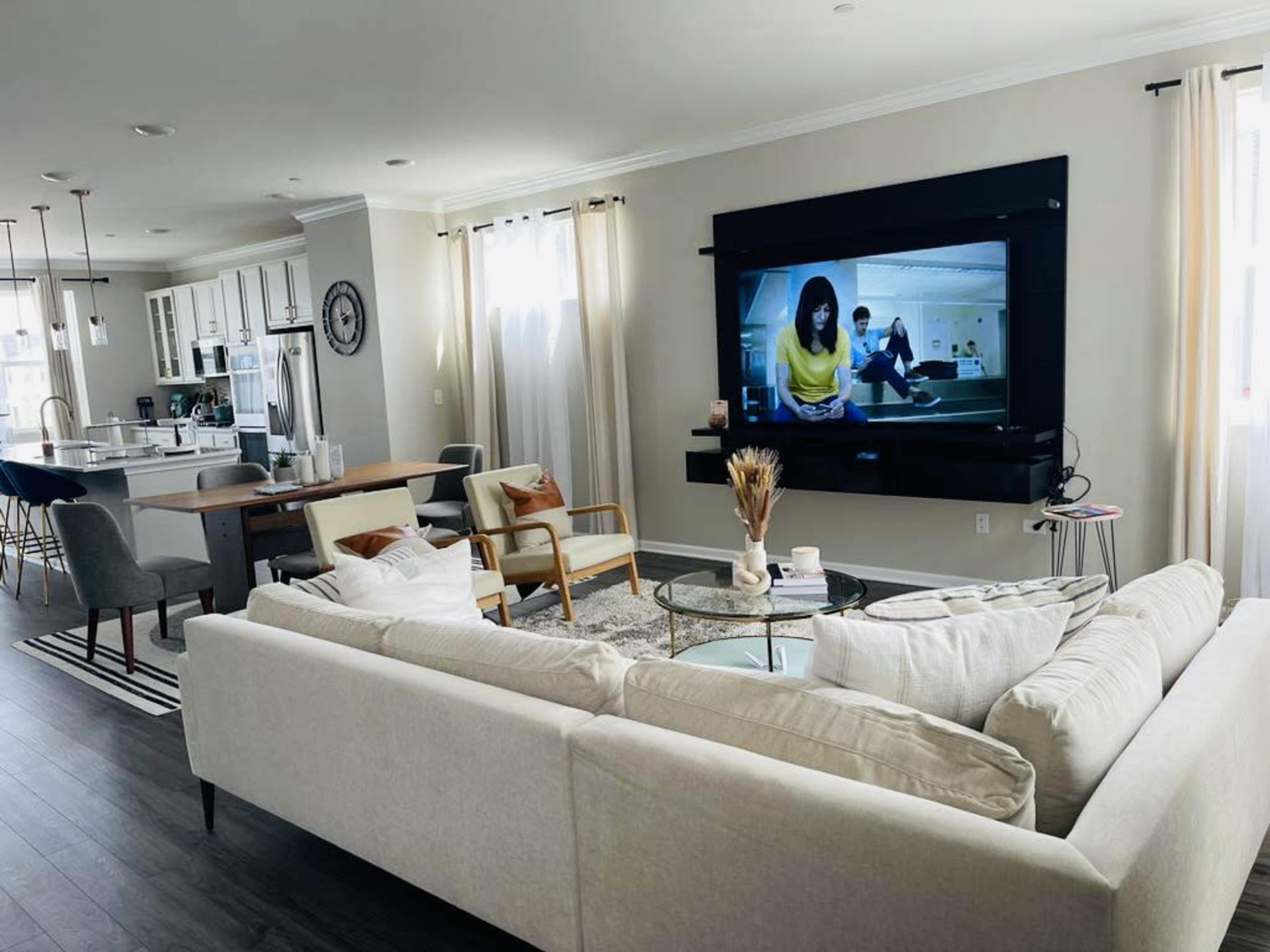 A modern living room features a large sectional sofa facing a mounted television, with a dining area and kitchen visible in the background.
