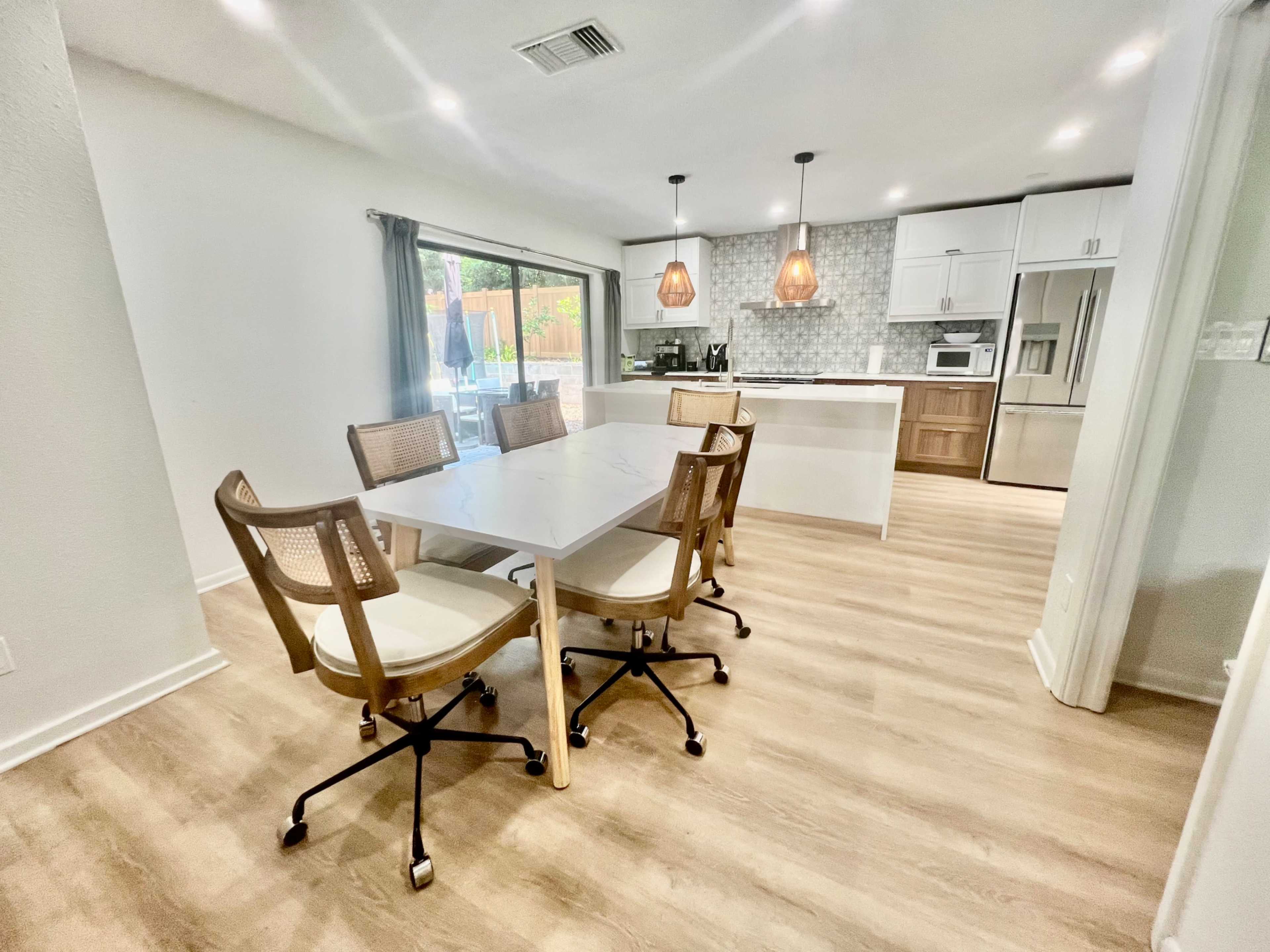 The image shows a modern dining area with a white table and four rolling chairs, adjacent to a kitchen featuring pendant lights and a tiled backsplash.