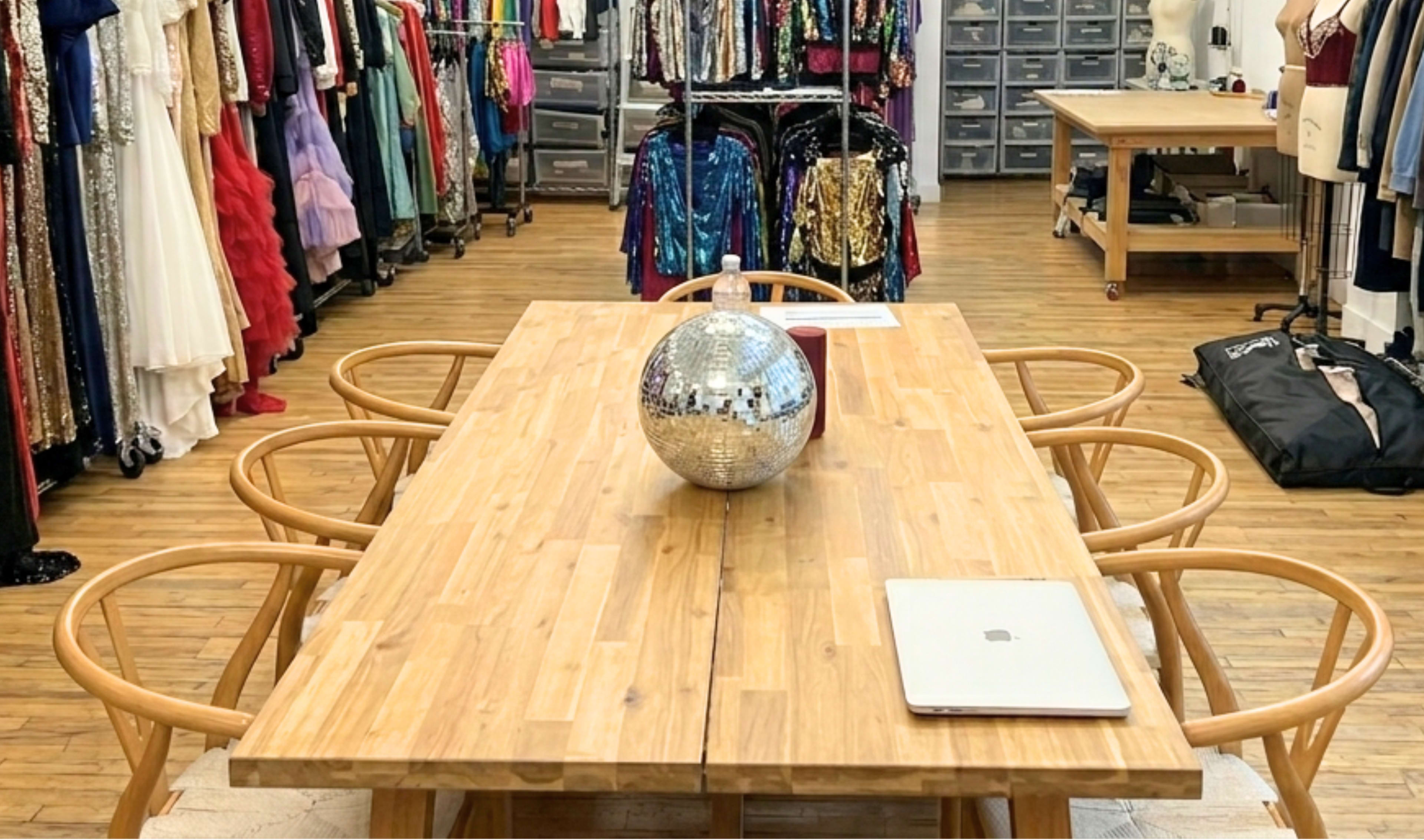 The image shows a wooden dining table with a disco ball centerpiece, surrounded by four chairs, in a boutique filled with colorful garments and storage units.
