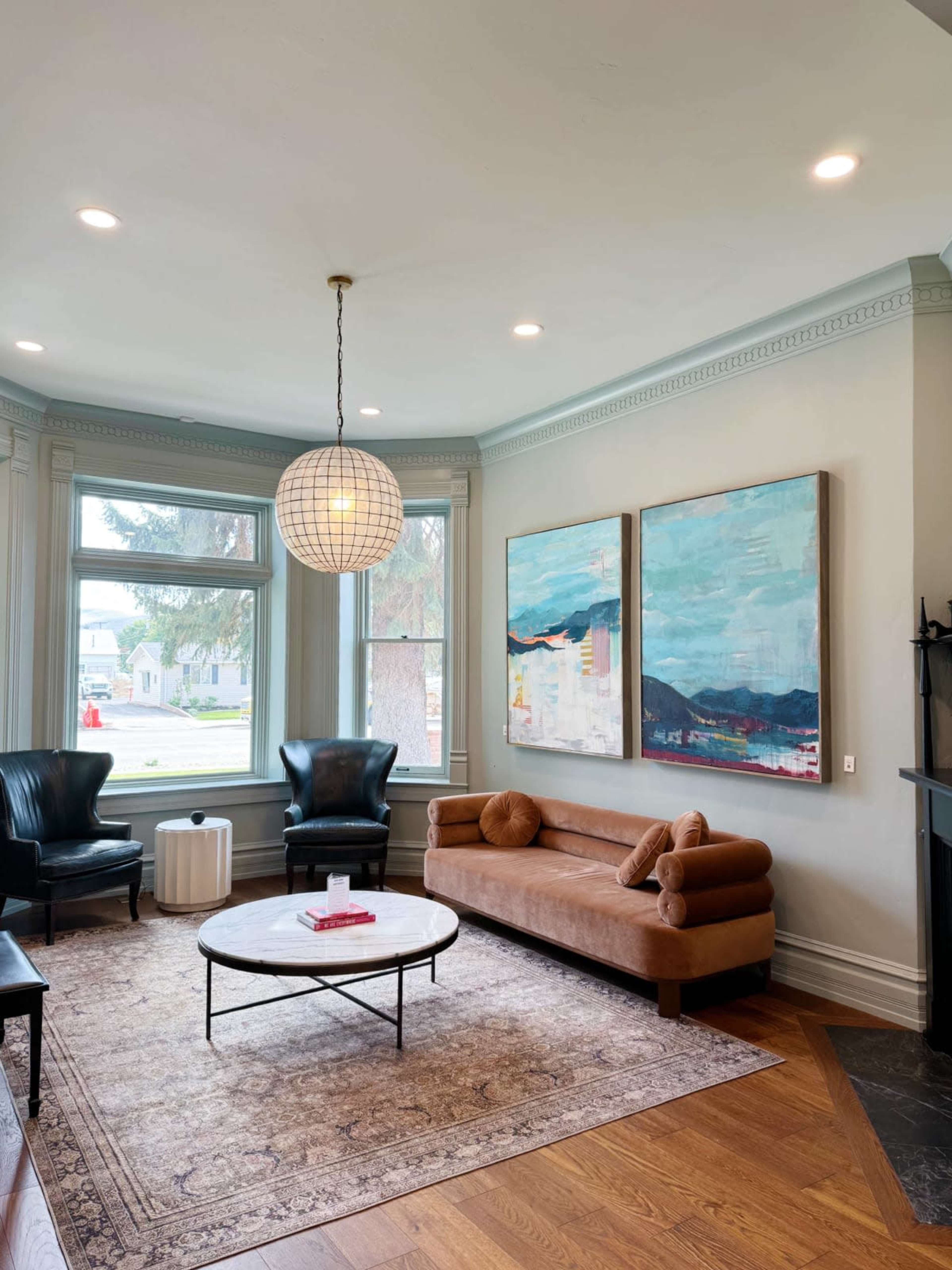 A living room with a tan leather sofa, two black armchairs, a round coffee table, and large windows featuring abstract artwork on the walls.
