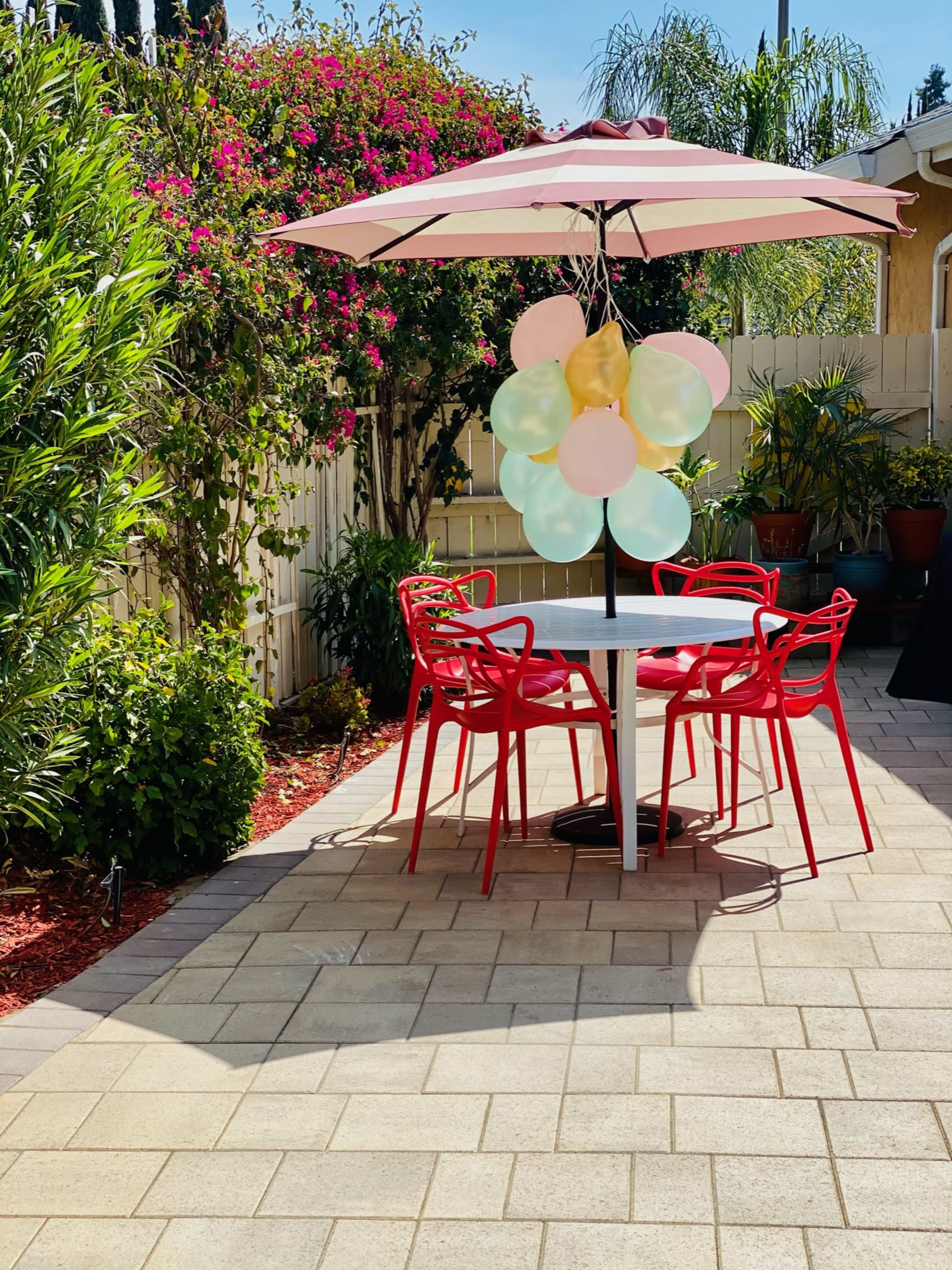 A shaded outdoor seating area features a white table surrounded by four red chairs and colorful balloons hanging from a large umbrella.