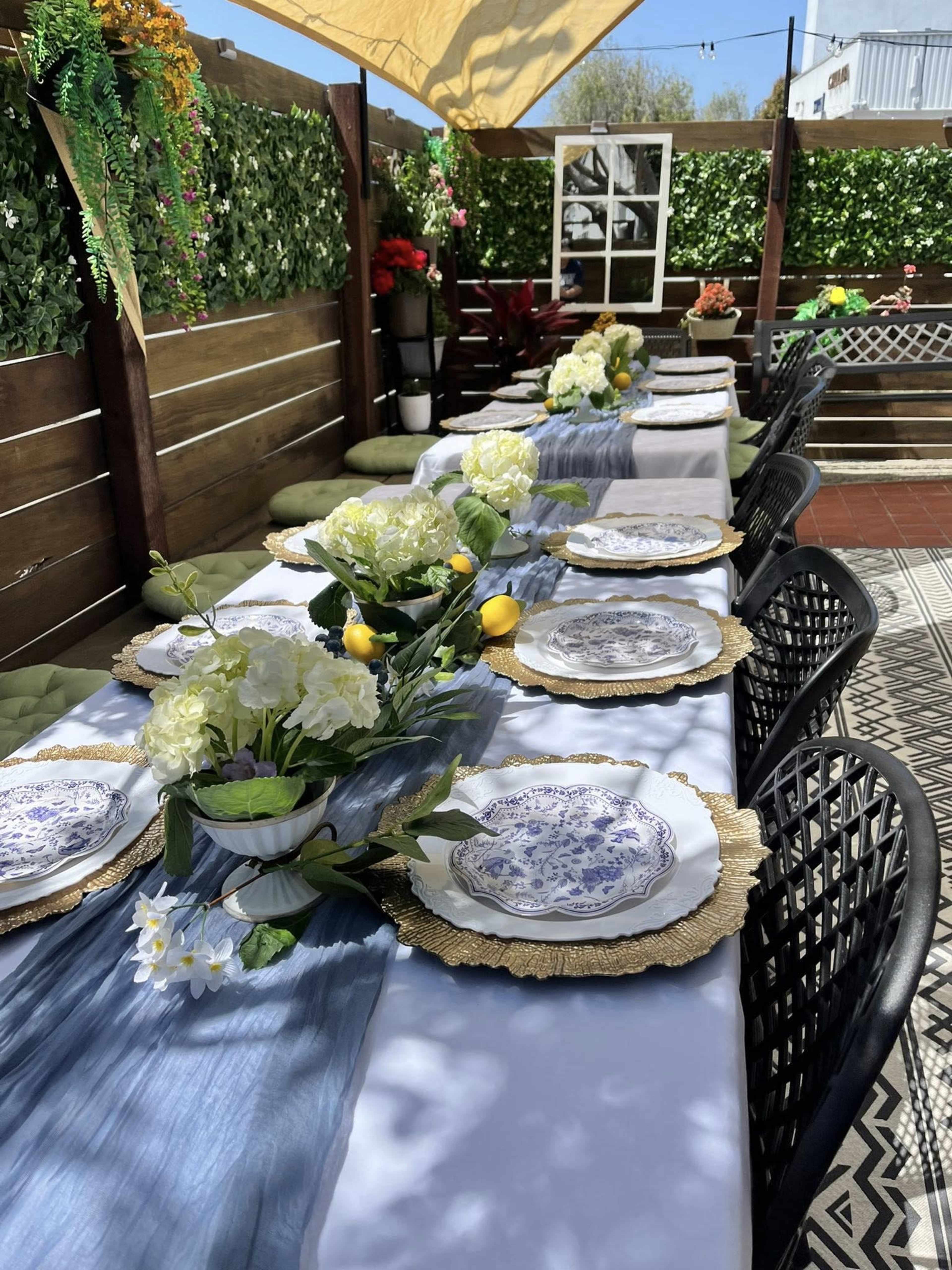 A long outdoor table is set with floral centerpieces, plates, and gold chargers, surrounded by black chairs and greenery.