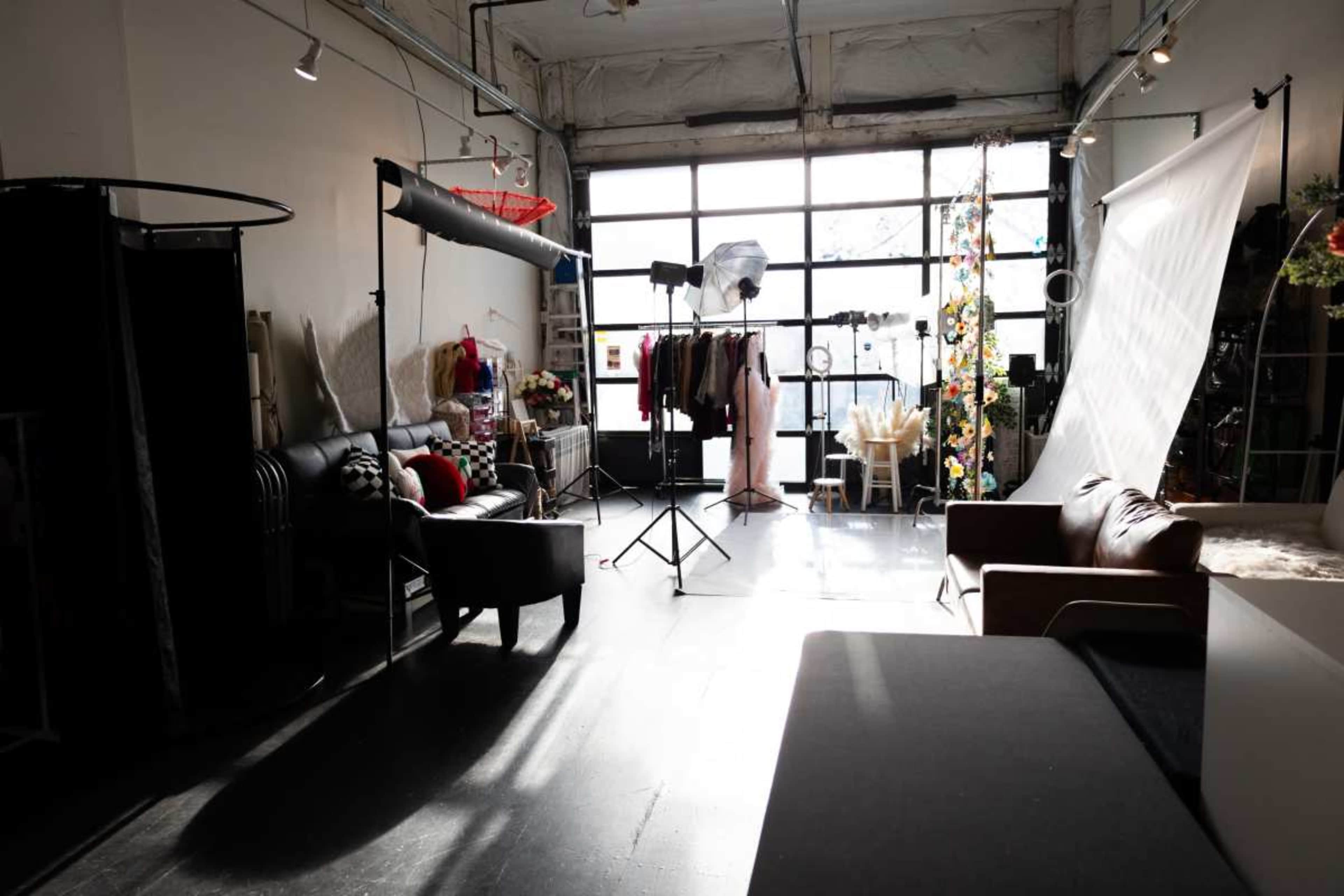 The image shows a photography studio with various props, clothing racks, and lighting equipment set up near large windows.
