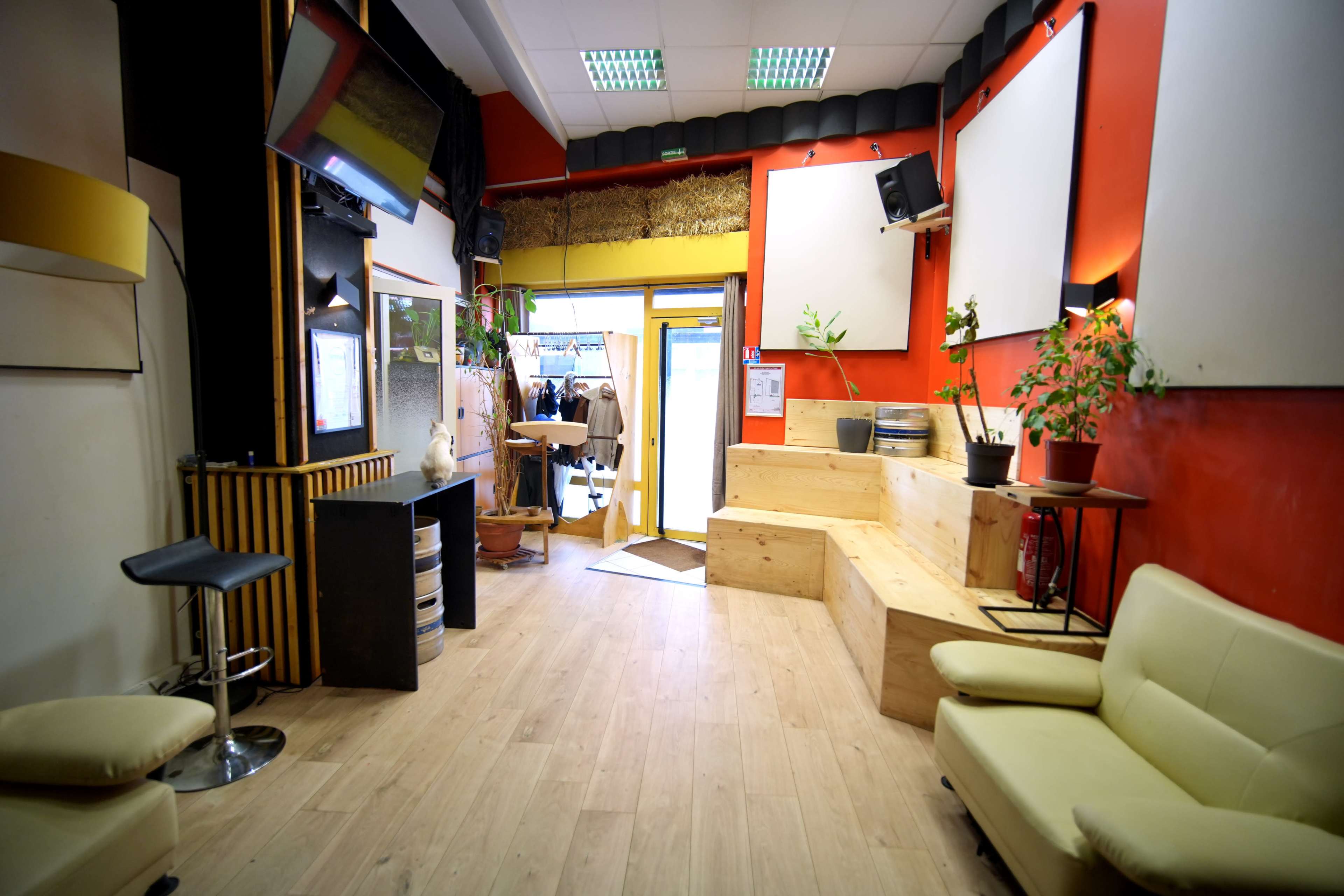 The interior of a spacious room features light wooden flooring, a small seating area, and an entrance leading outside, with various plants and decorative elements along the walls.