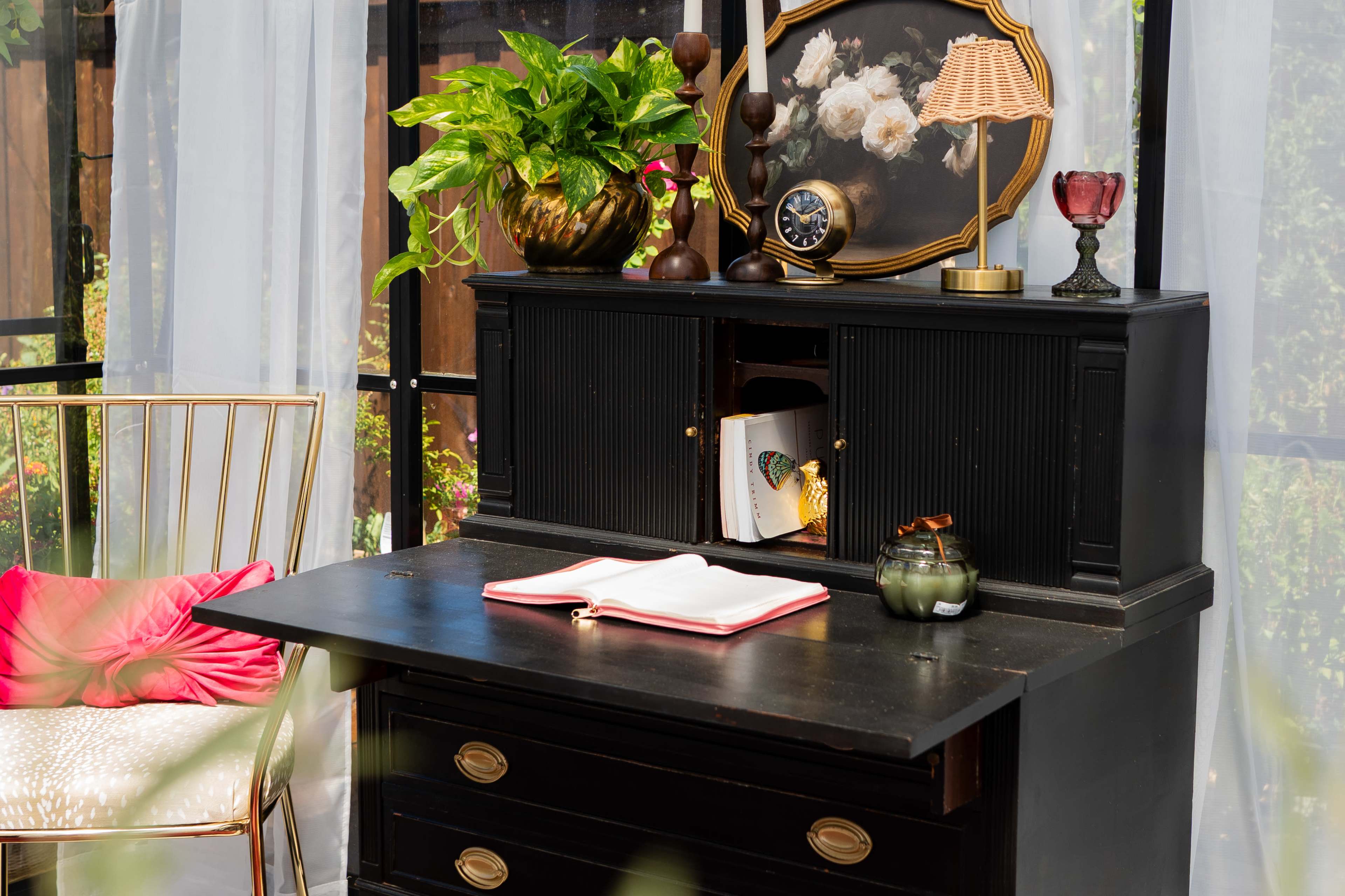 A black desk with a textured surface is positioned against a backdrop of sheer curtains, adorned with a potted plant, a vintage clock, and an open book.