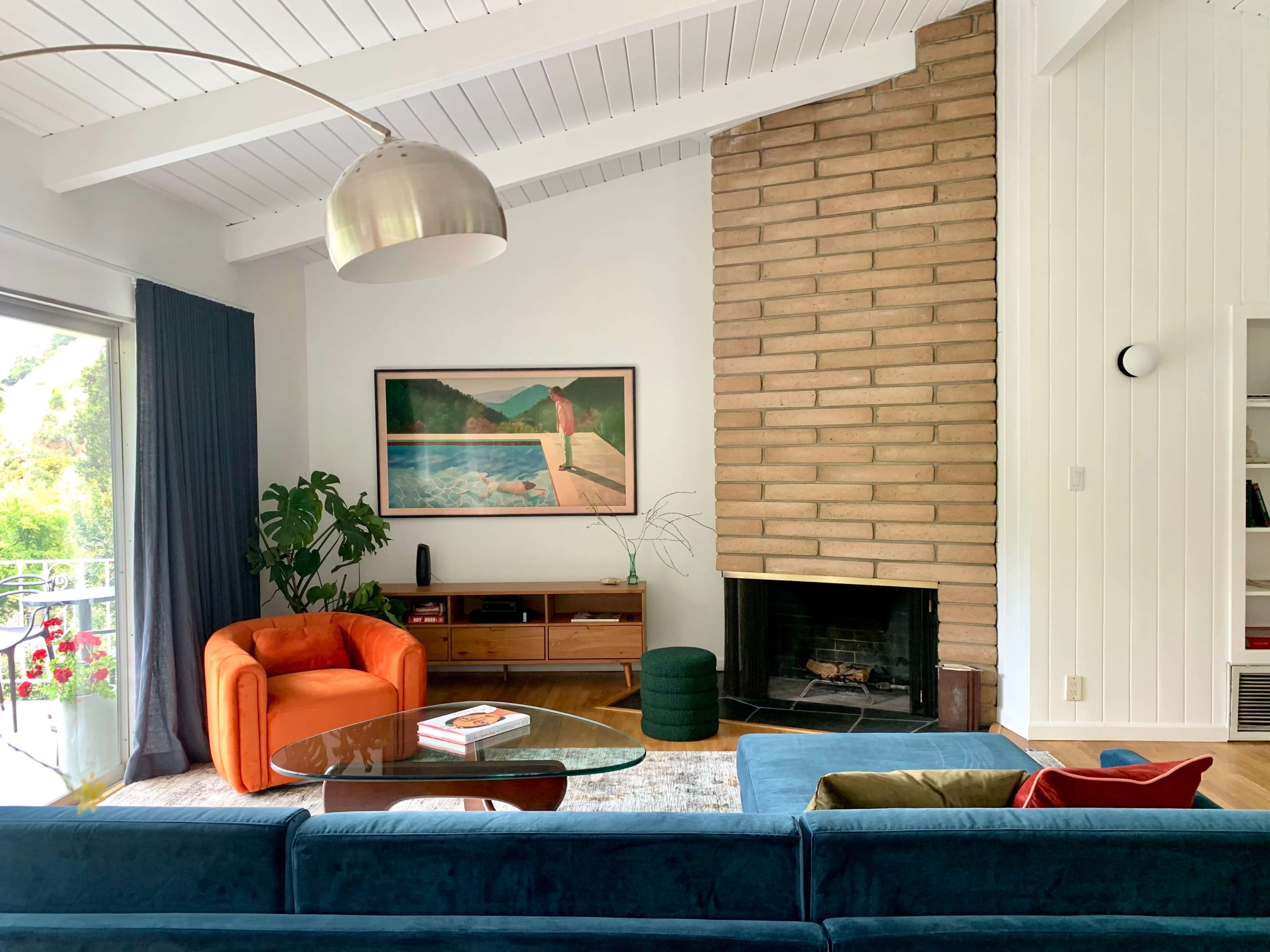 A living room features a brick fireplace, a large window with drapes, and a mix of colorful furniture, including an orange armchair and a blue sofa.