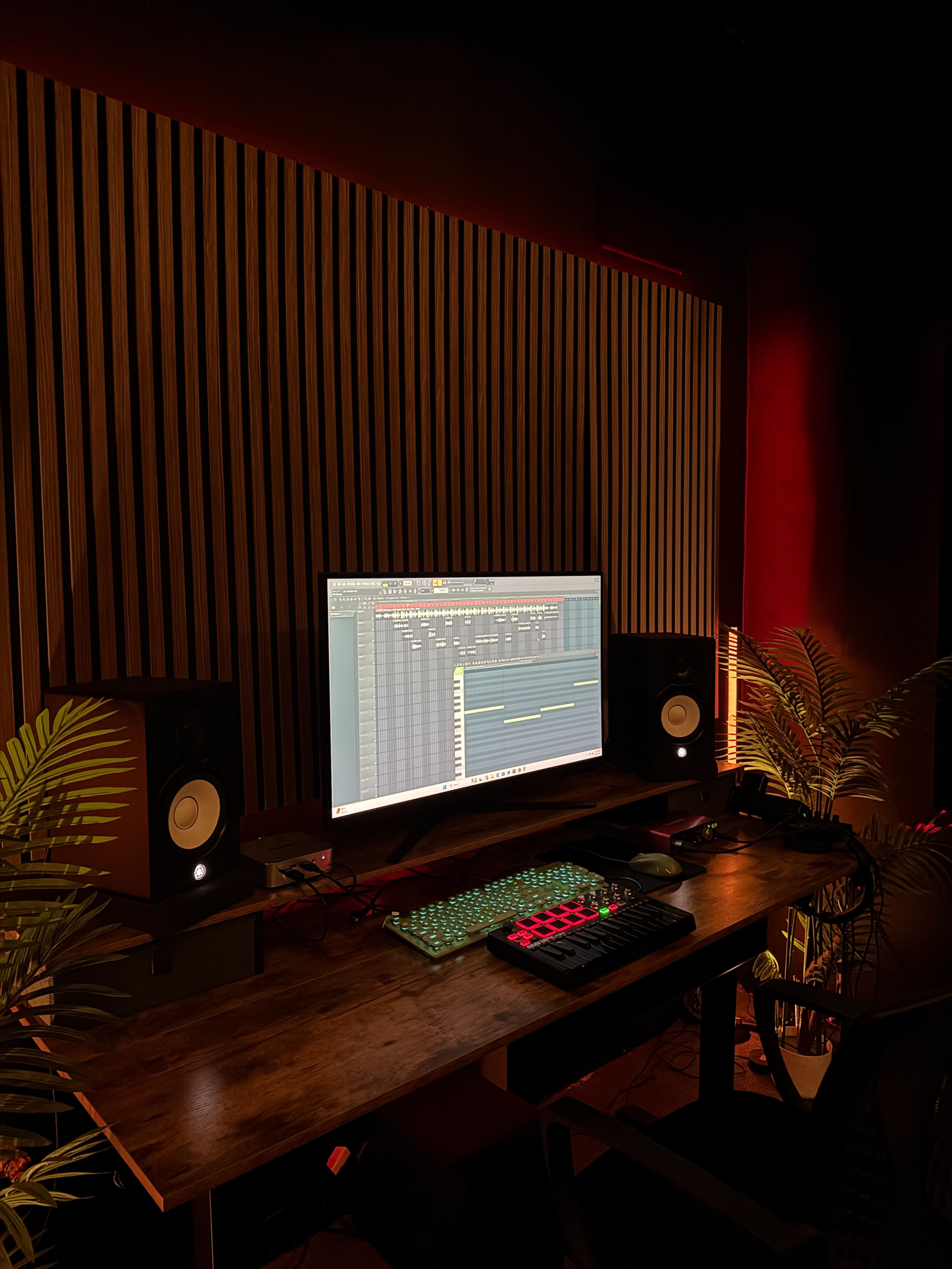 The image shows a dimly lit music production studio with a large monitor, two speakers, and a keyboard on a wooden desk, surrounded by plants and a slatted wall.