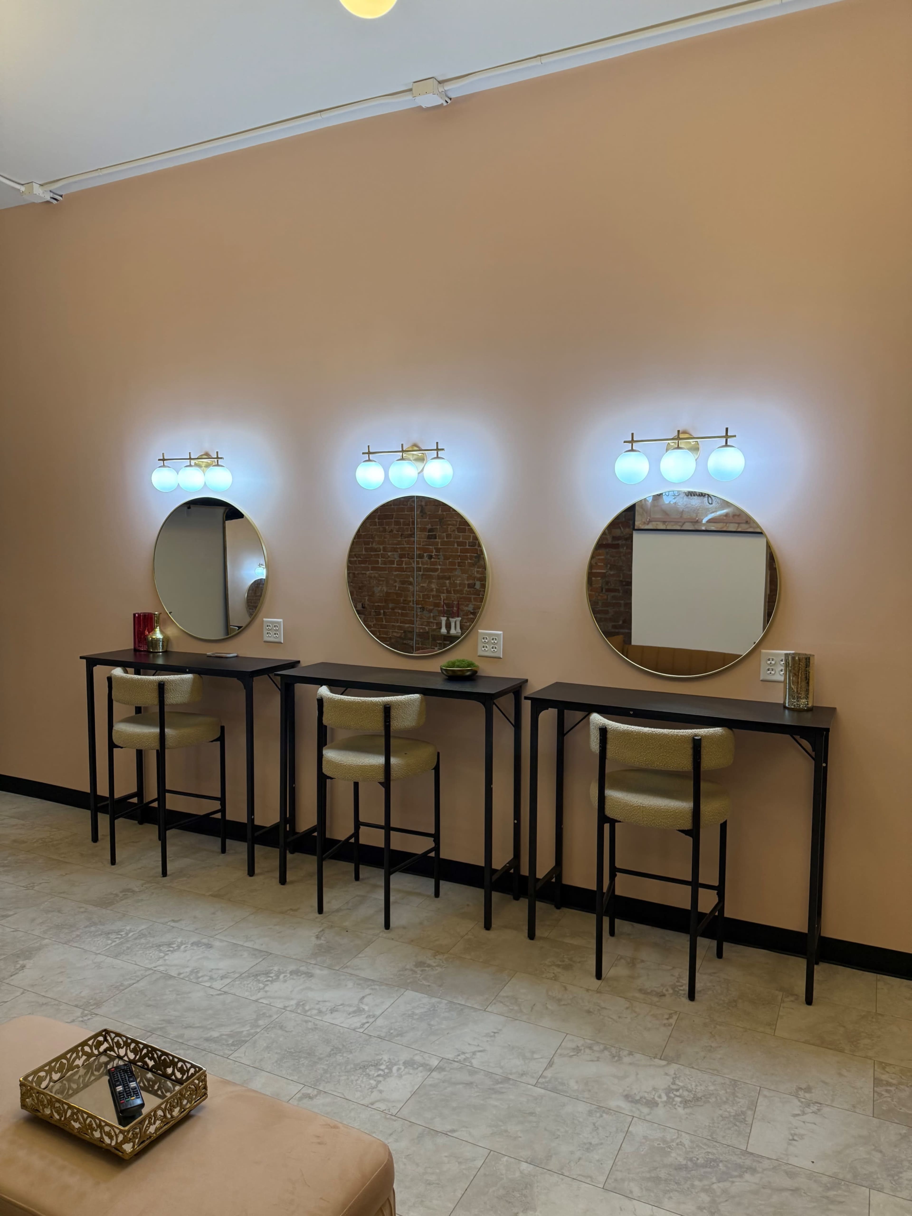 The image shows a stylish vanity area with three round mirrors, each illuminated by wall-mounted lights, above minimalist black tables and beige stools against a light pink wall.