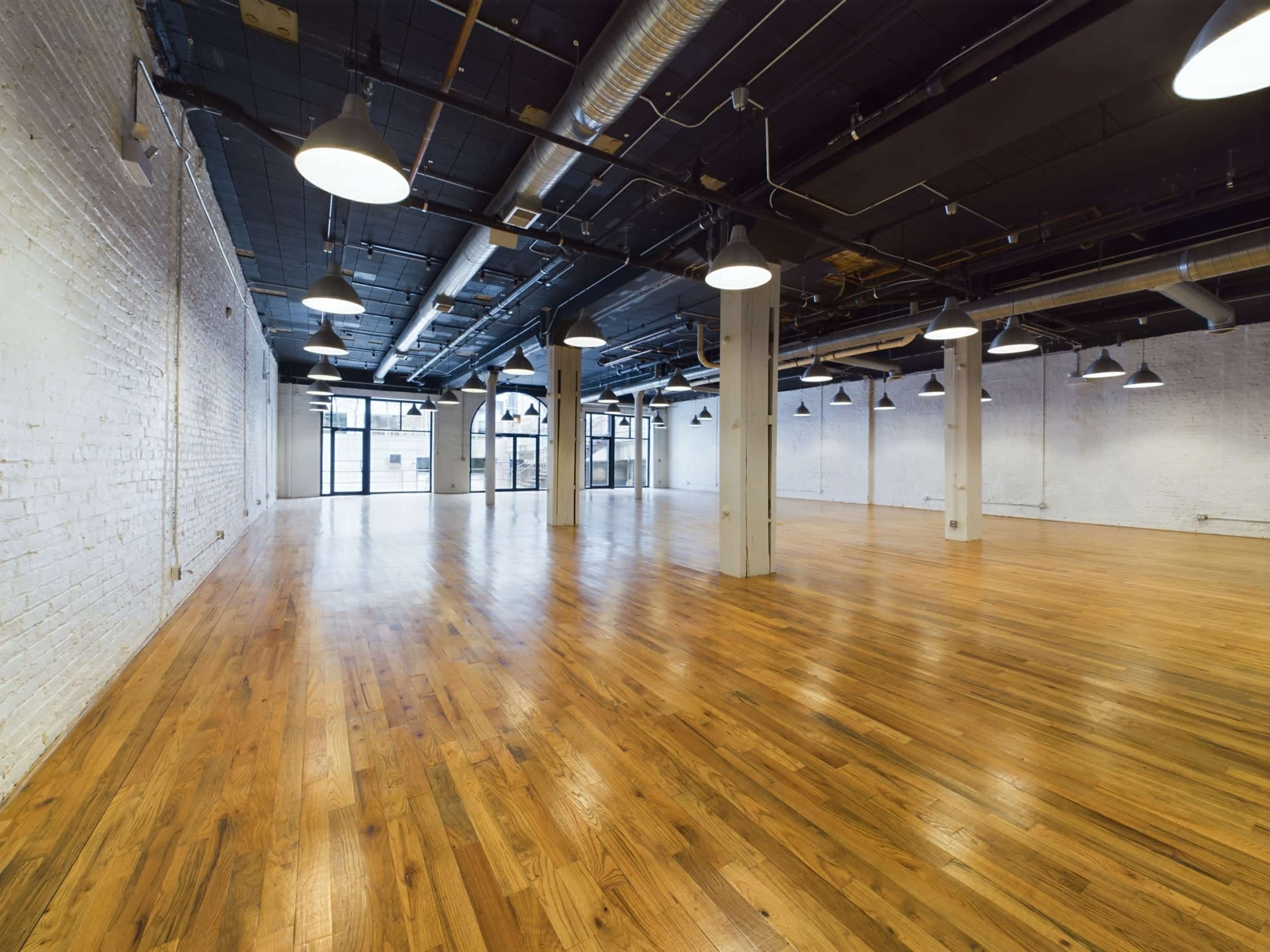 A spacious, empty room with polished wooden floors, high ceilings, and large windows letting in natural light.