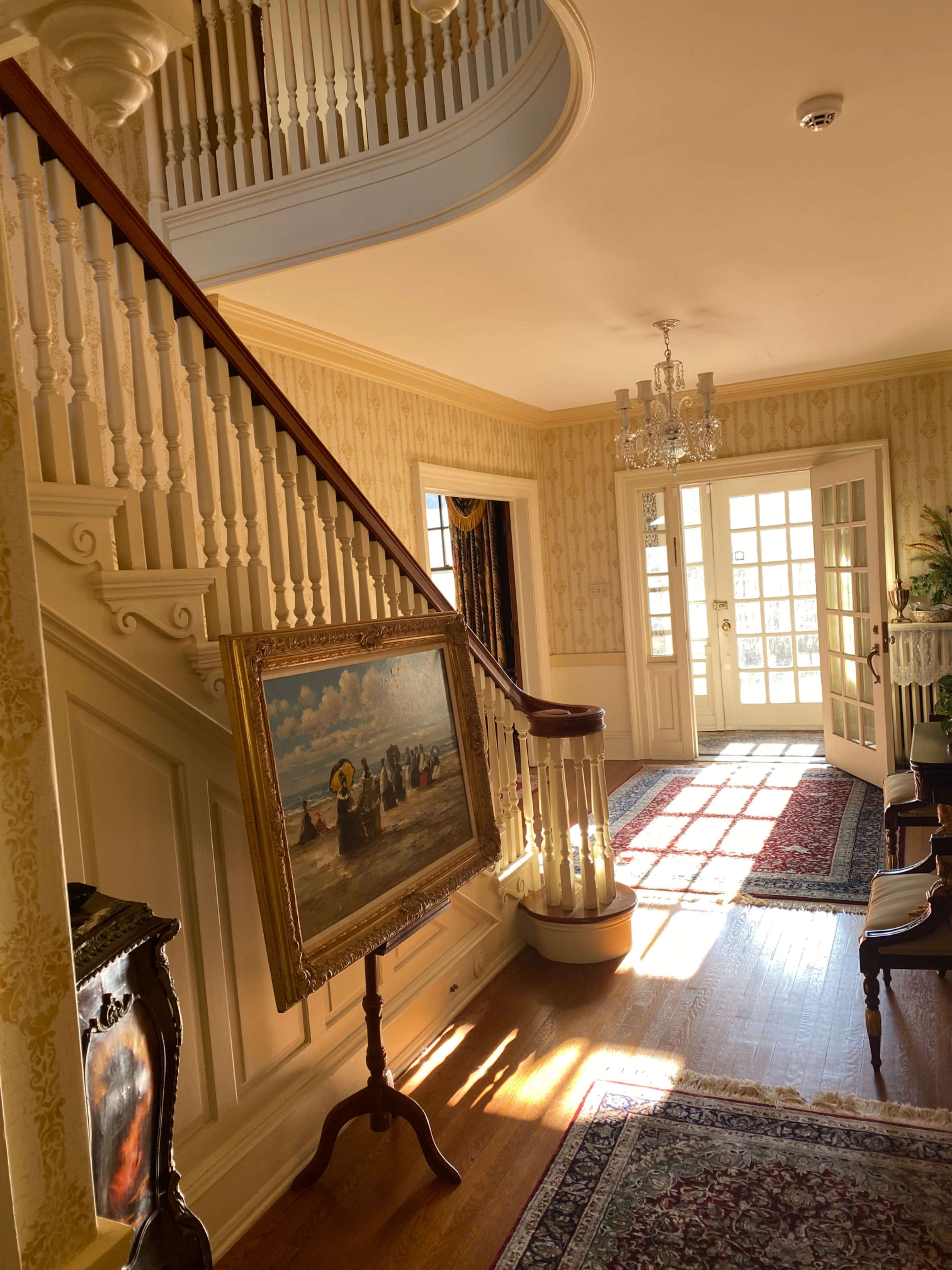A spacious hallway features a curved staircase, a large painting on an easel, and a chandelier, with sunlight illuminating the wooden floor.