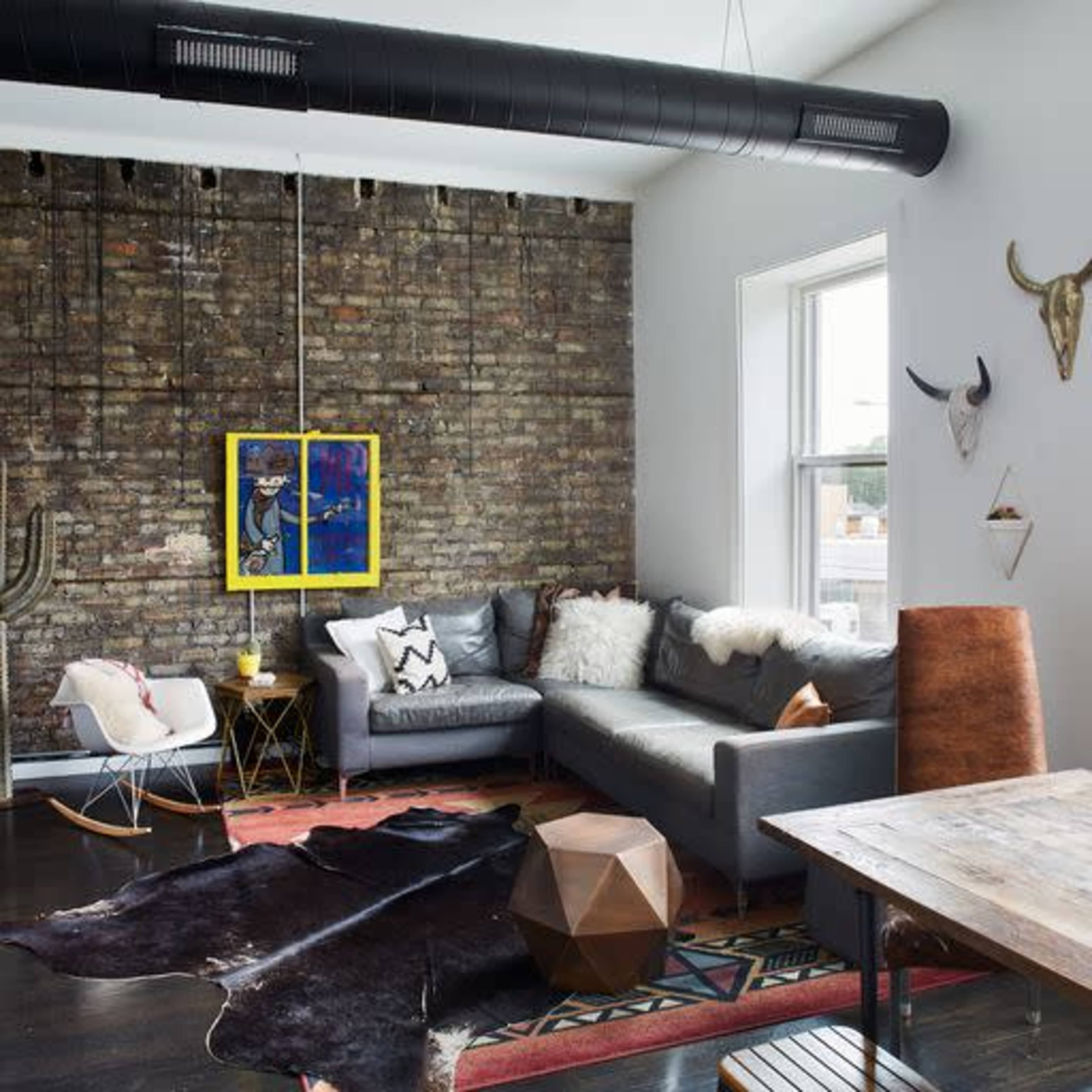 The scene features a modern living space with a gray sofa, an eclectic mix of decorations, and a large brick wall as a backdrop.