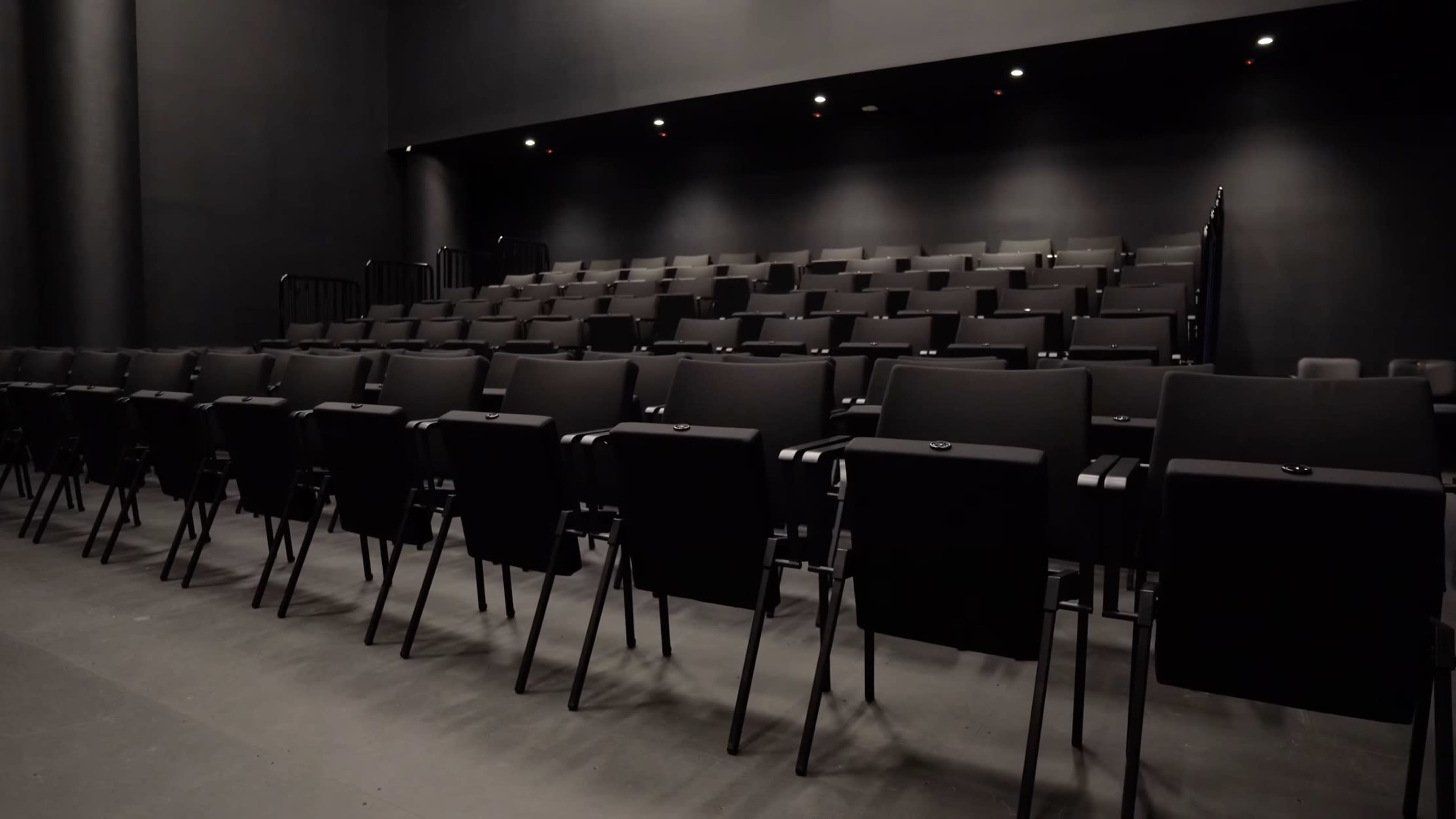 A row of black chairs arranged in a tiered seating layout in a dimly lit theater.