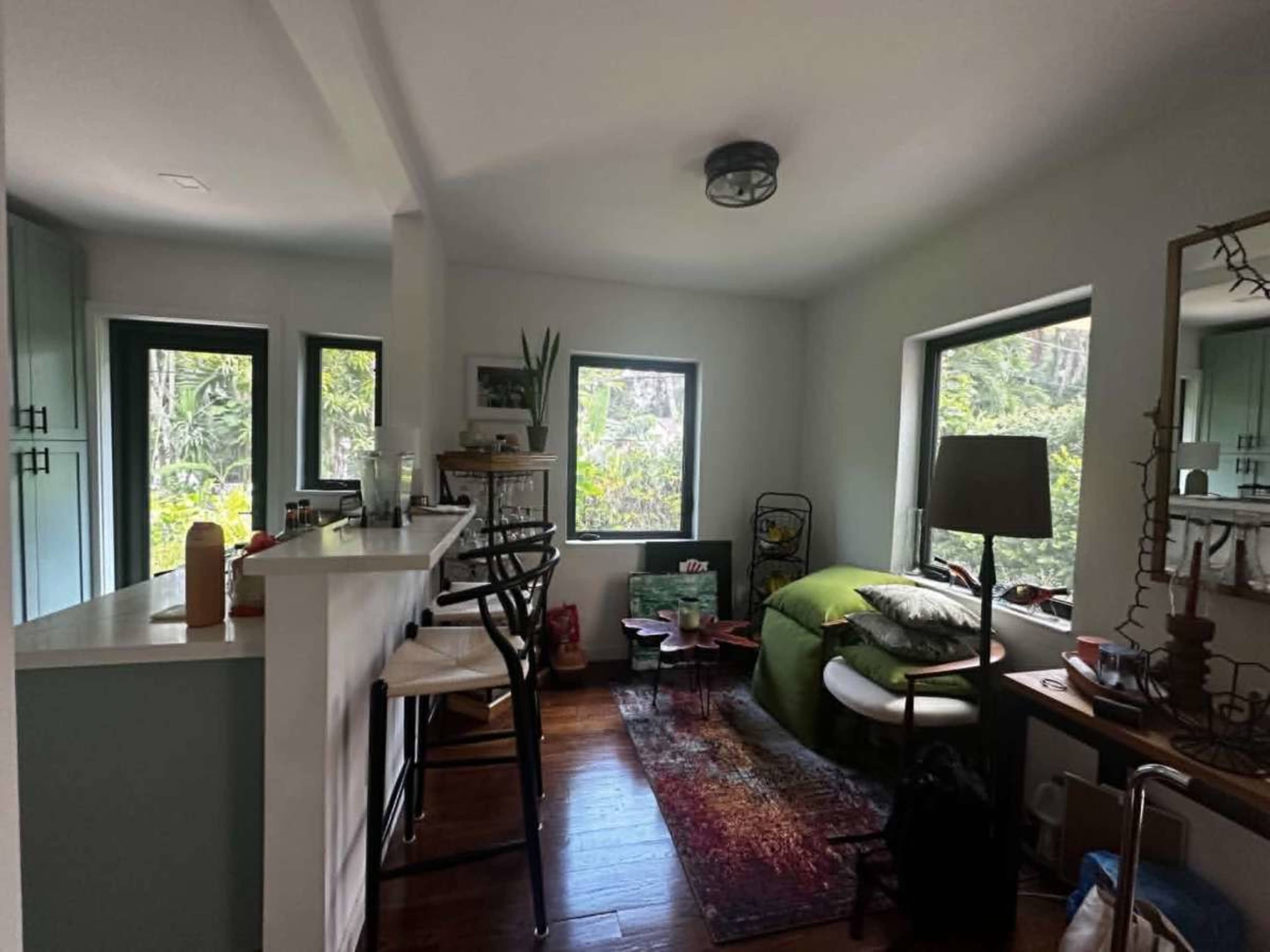 The image shows a cozy interior space featuring a bar counter with high stools, a green bed, and several windows overlooking a garden.