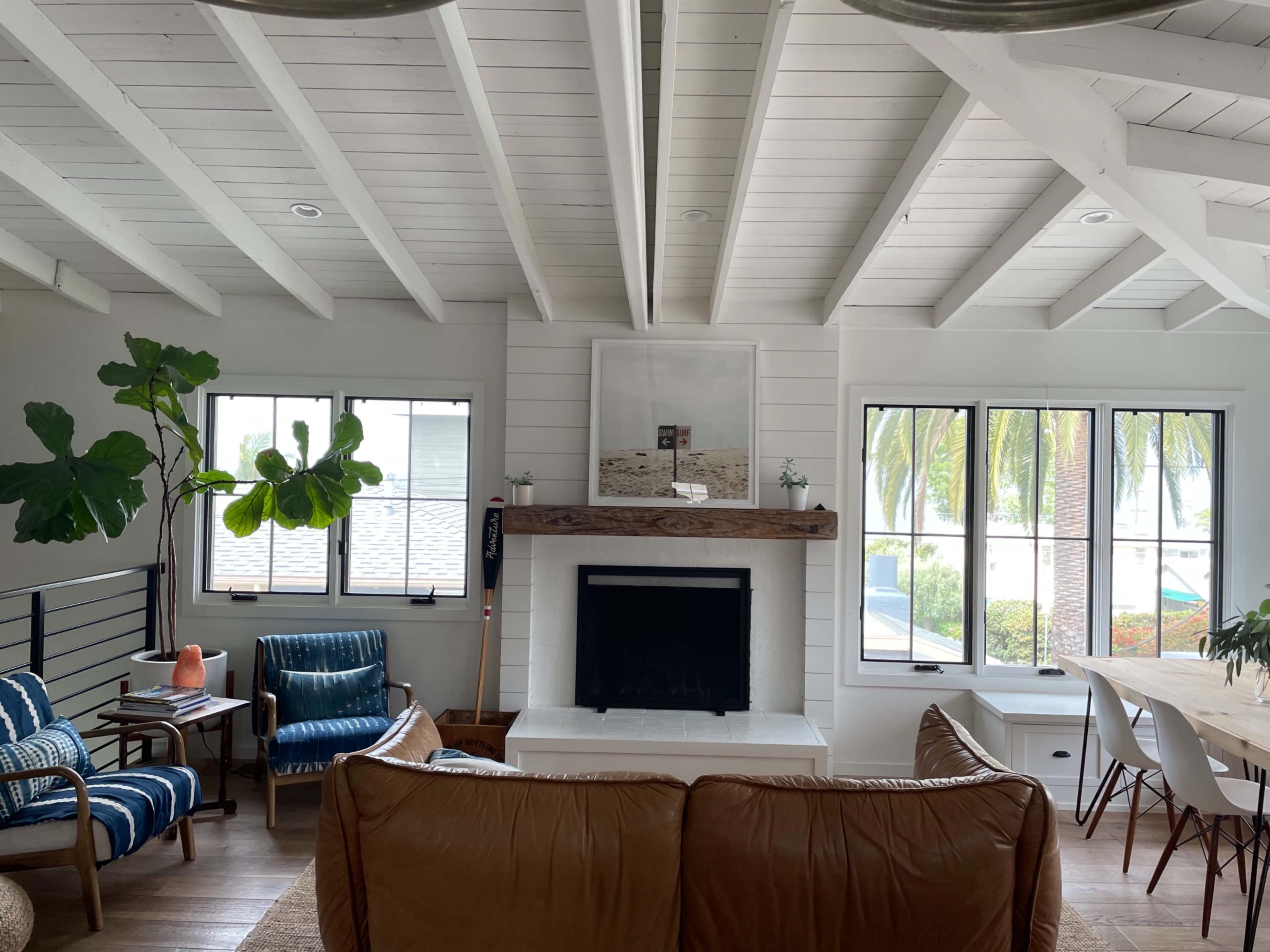 The image shows a spacious living room with a white-beamed ceiling, a fireplace with a wooden mantel, and large windows overlooking palm trees and a dining area.