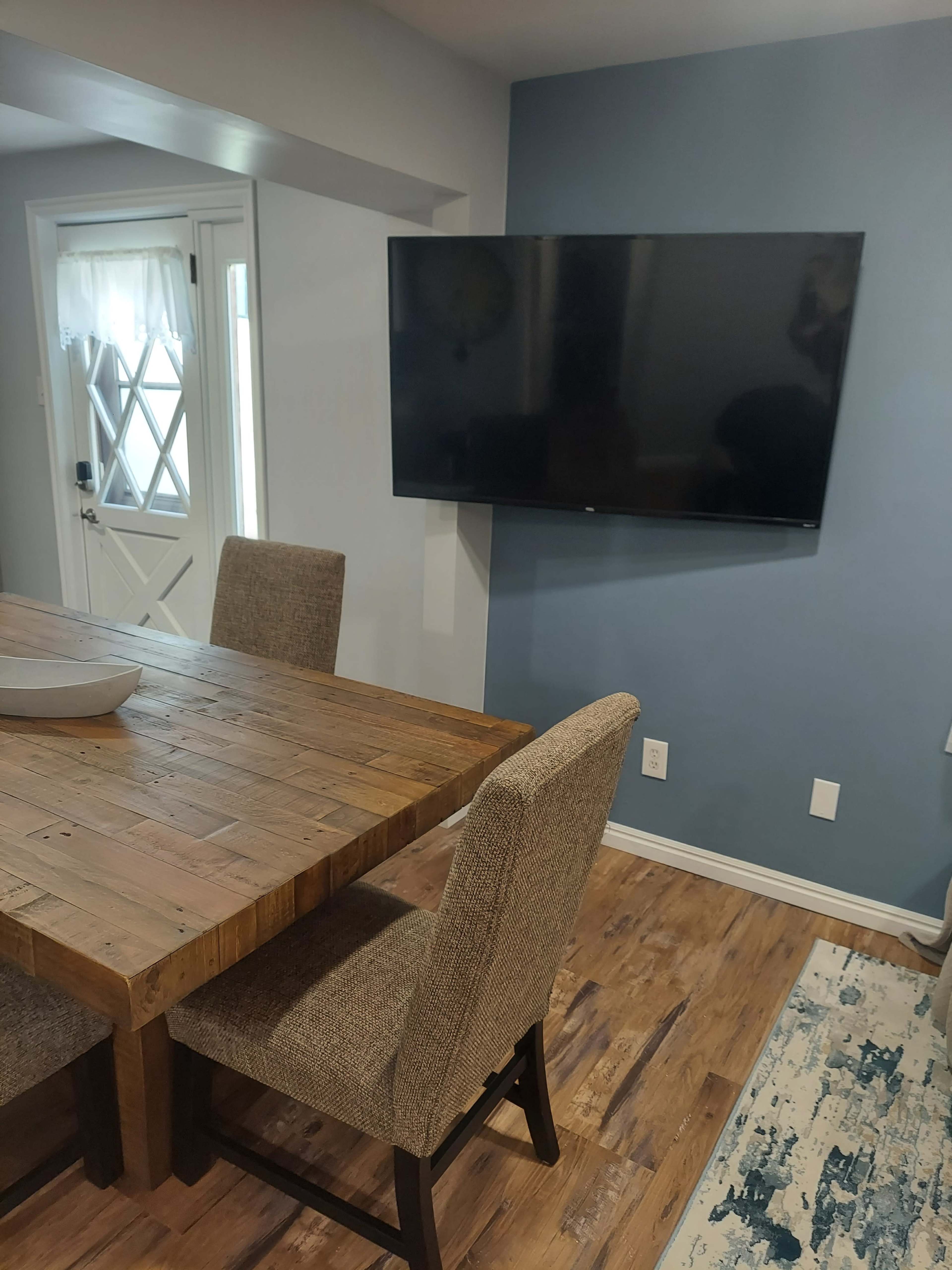 A wooden dining table with two upholstered chairs is positioned in a room featuring a mounted TV on the wall and a door leading outside.