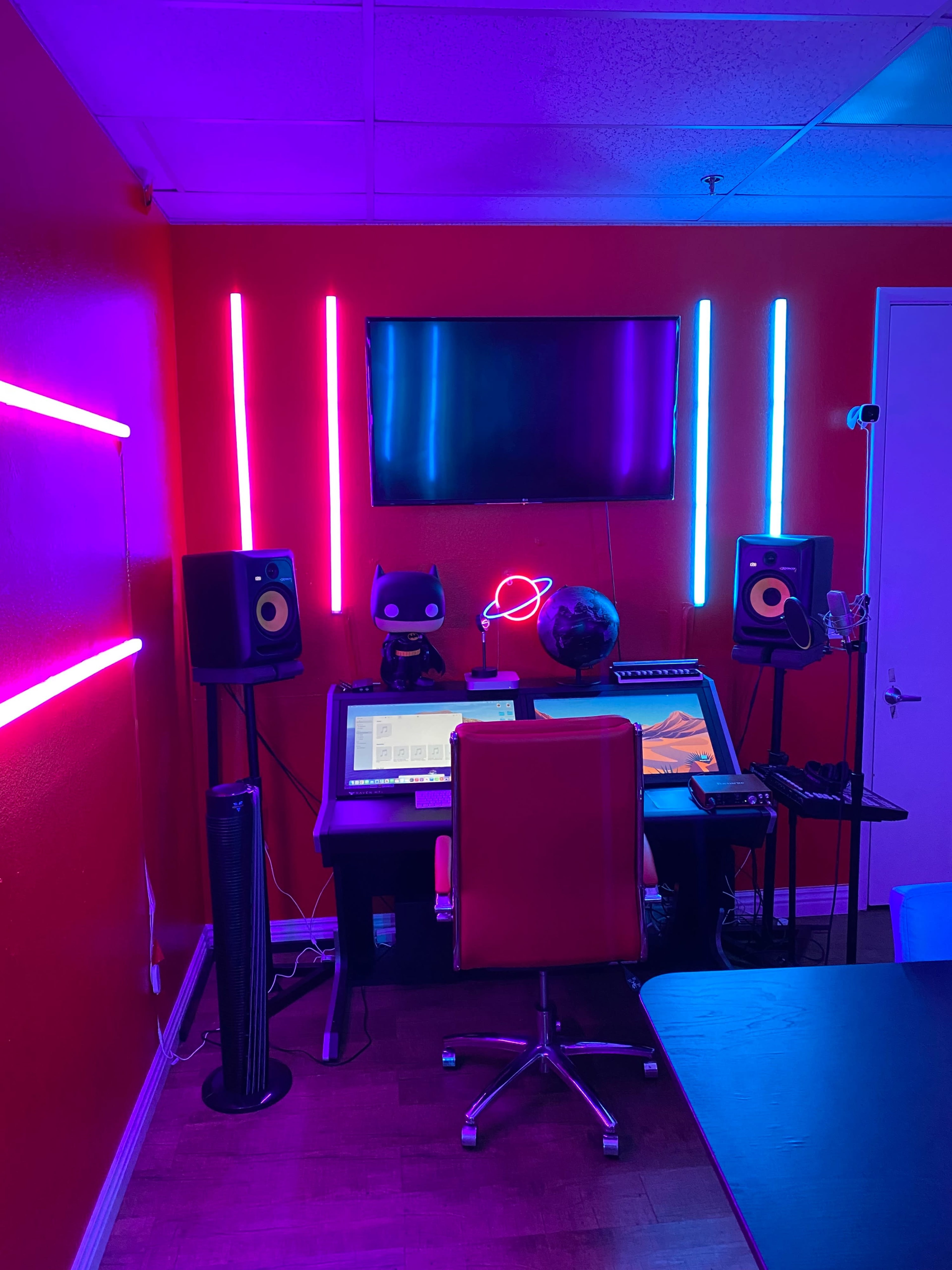 A room with red walls, featuring a desk with a computer setup, speakers, a globe, and neon lights in blue and purple.
