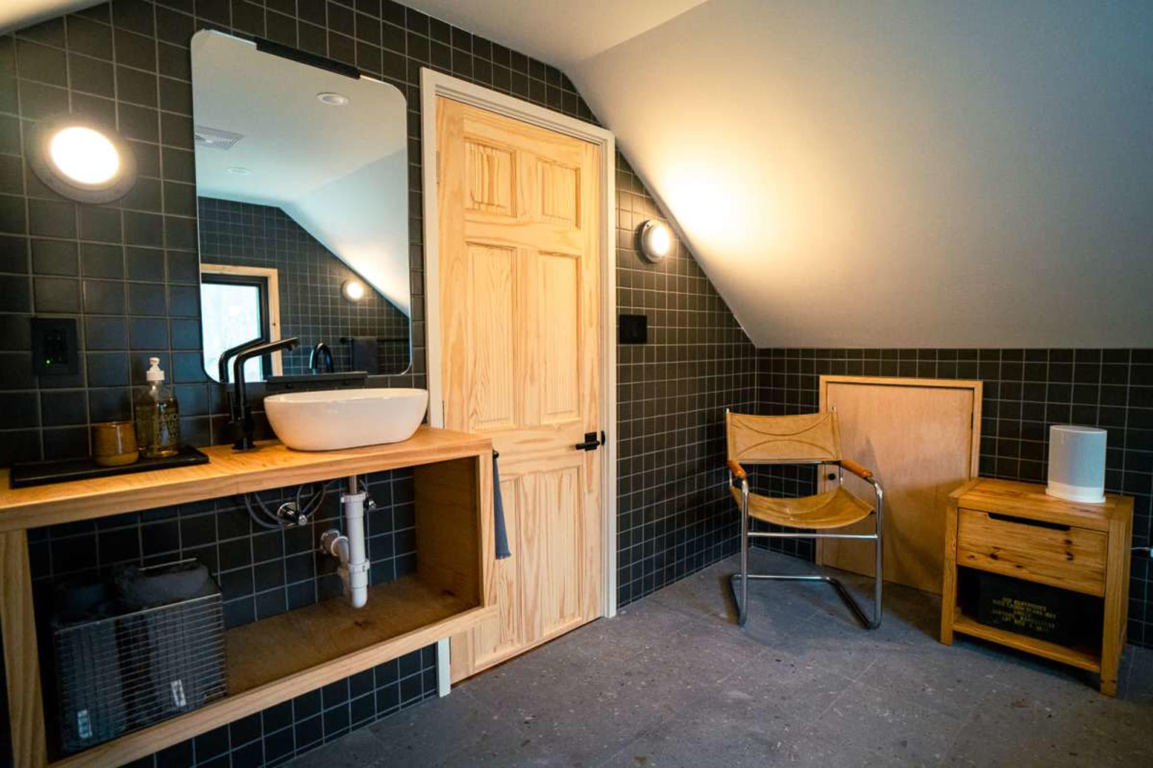 The image shows a modern bathroom featuring a wall-mounted mirror, a sink on a wooden shelf, and a corner with a chair and nightstand.