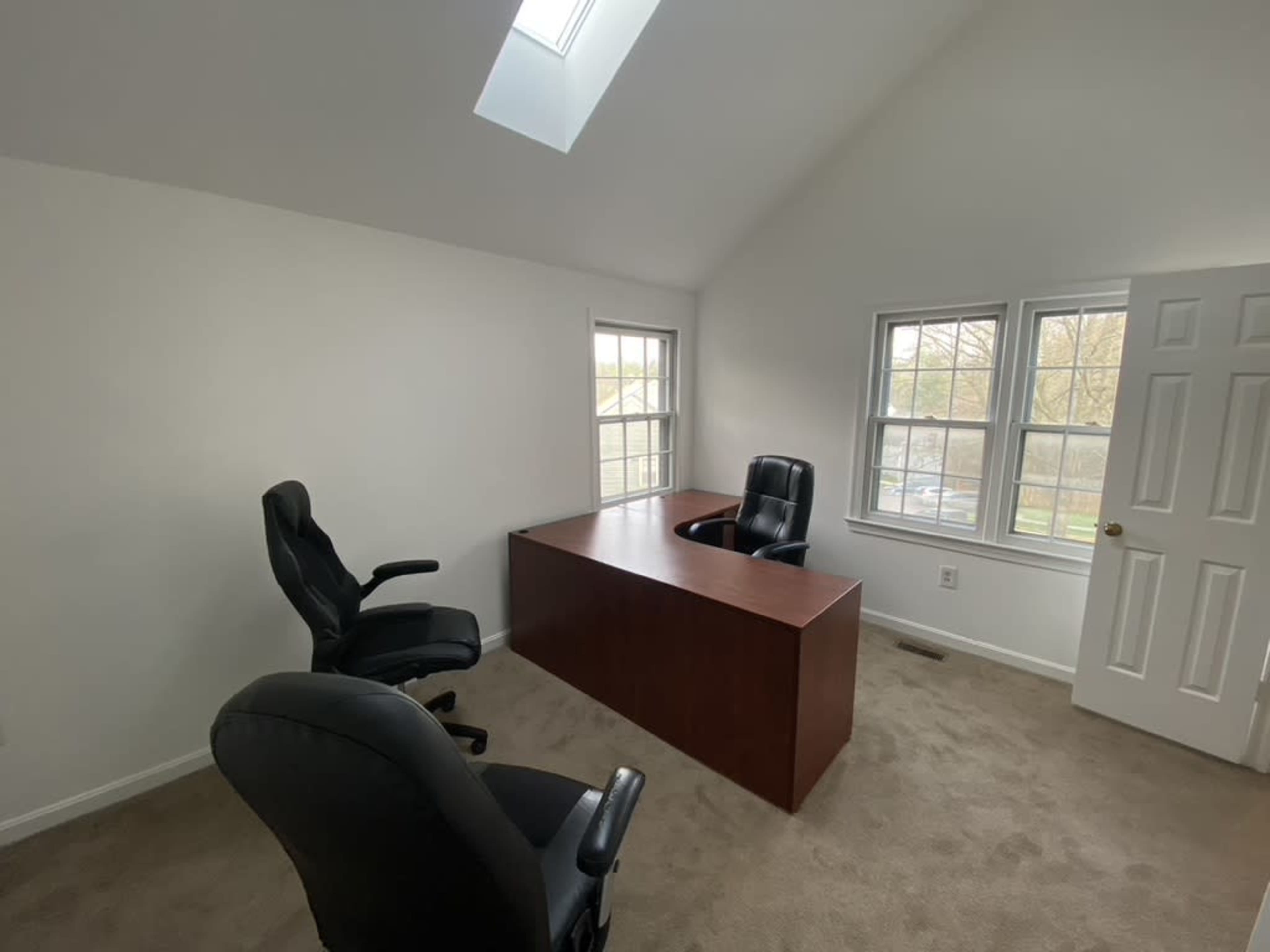 An empty office features a wooden desk and two black chairs, with windows allowing natural light to enter.
