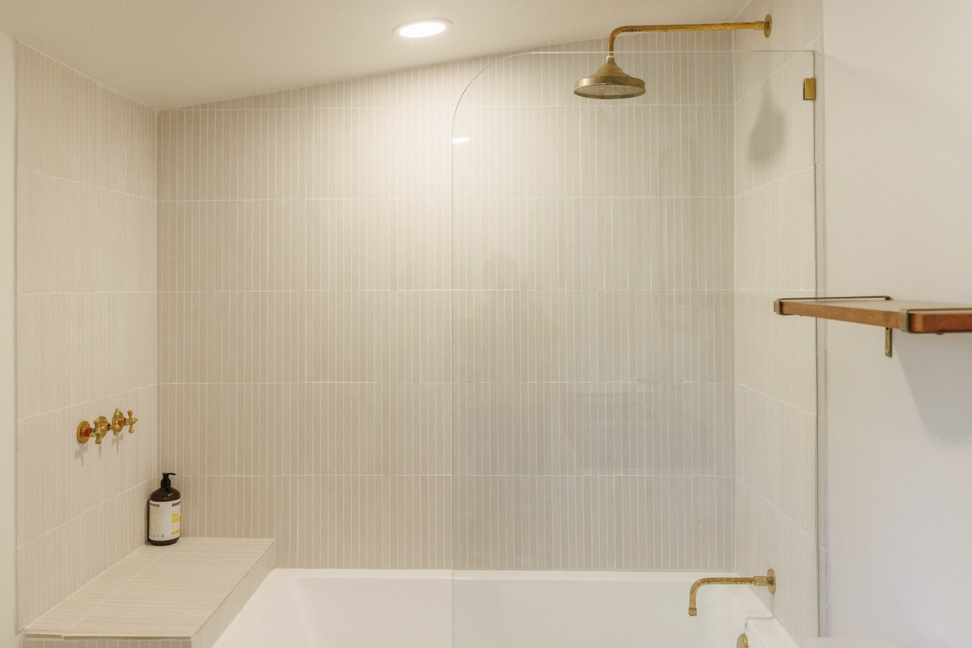 A minimalist bathroom with a shower area featuring a glass partition and brass fixtures, alongside a bathtub and a shelf against a light-colored tiled wall.