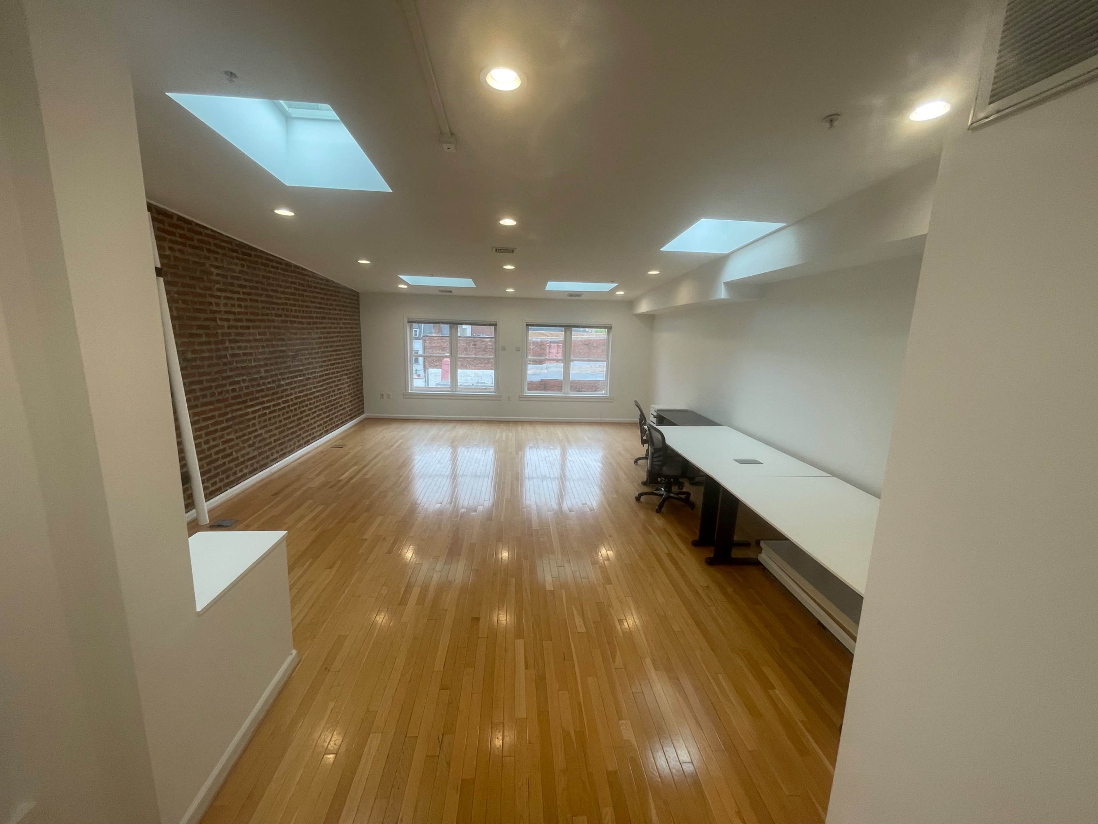 The image shows a spacious, modern room with hardwood floors, a brick accent wall, and several skylights allowing natural light to enter, along with a long table and a chair against one wall.