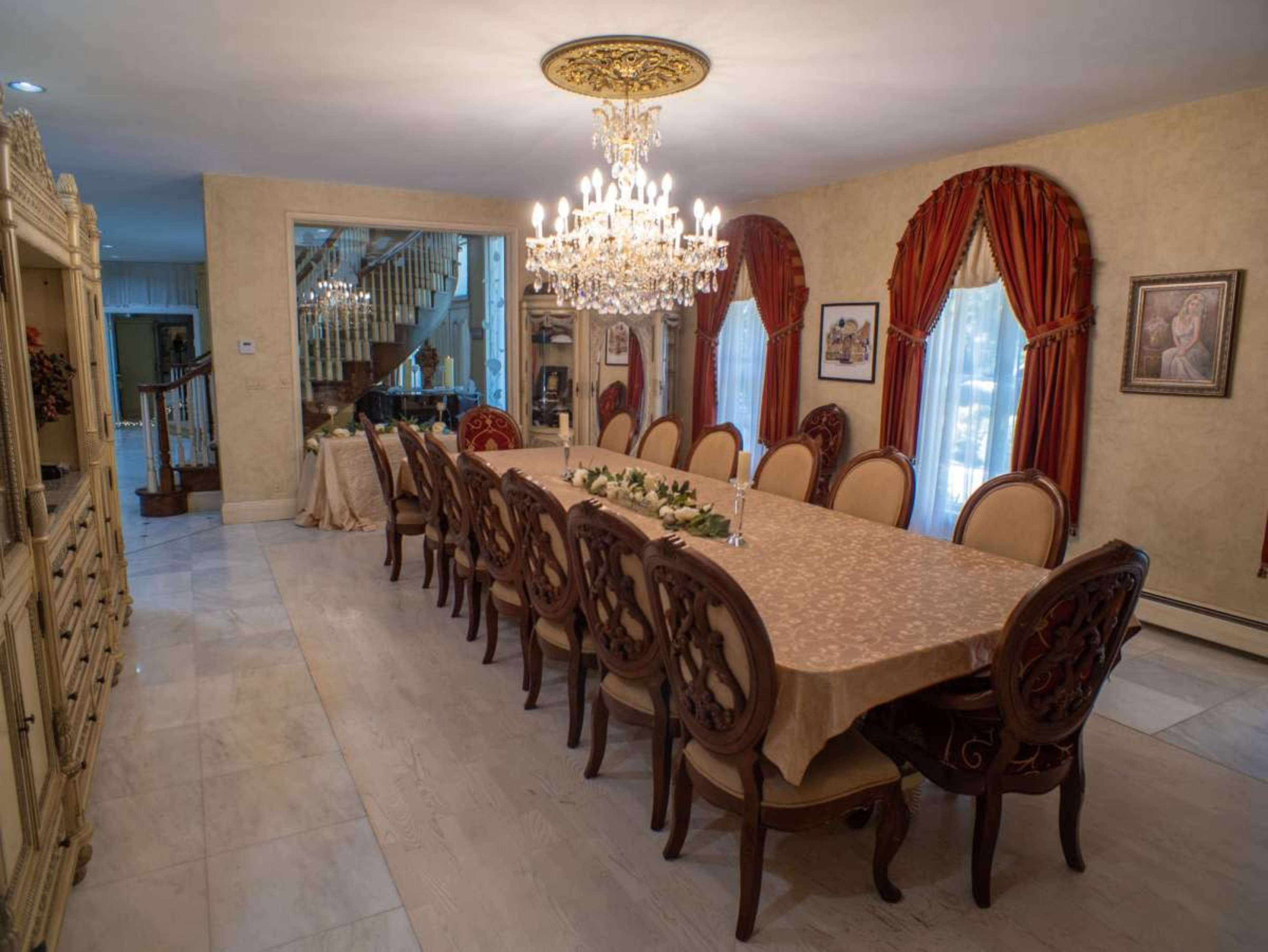 A long dining table with ornate chairs is set in a spacious room adorned with a chandelier and decorative drapes.