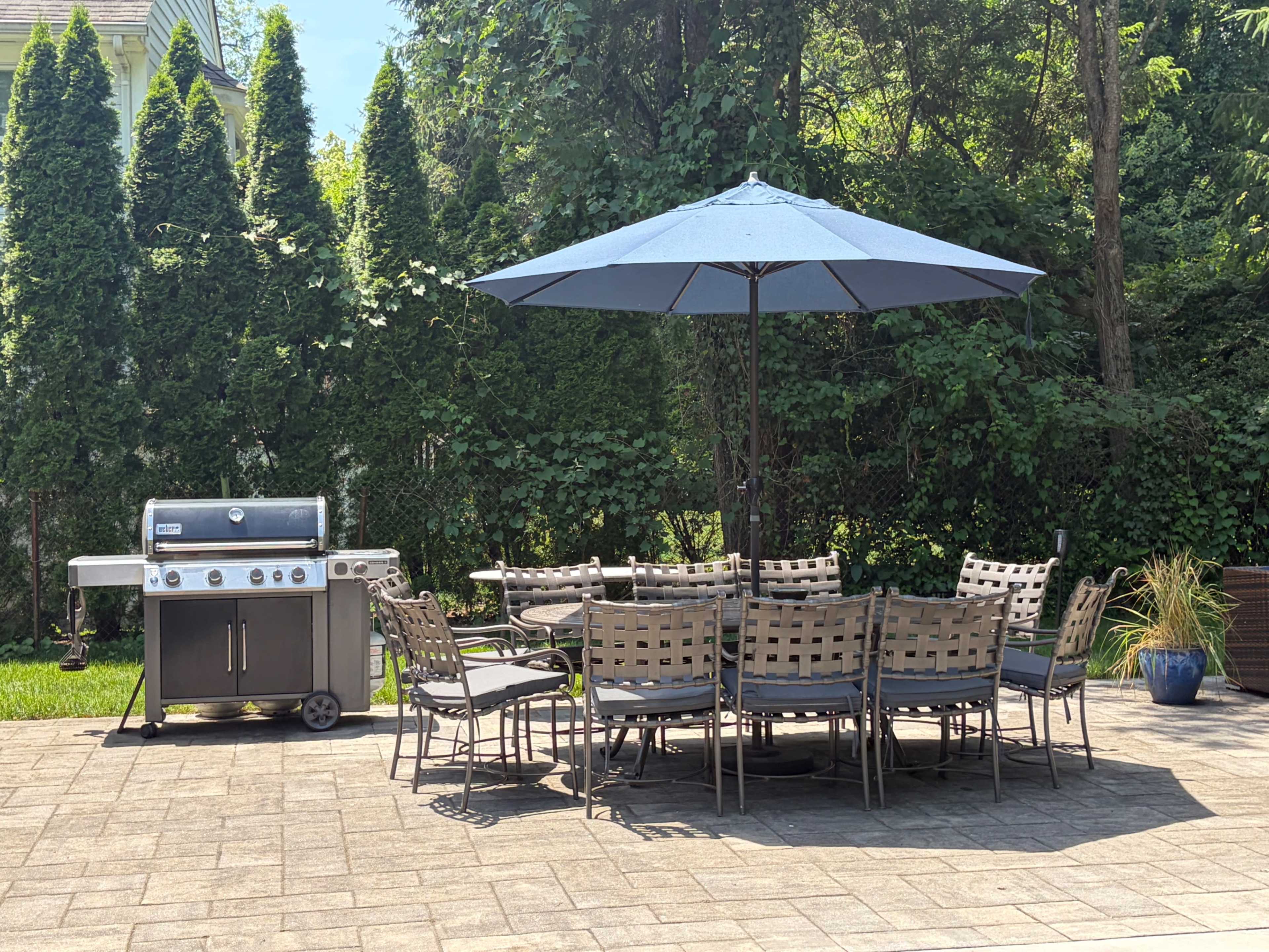 A patio area features a large dining table with multiple chairs, a blue umbrella, and a barbecue grill, surrounded by greenery.