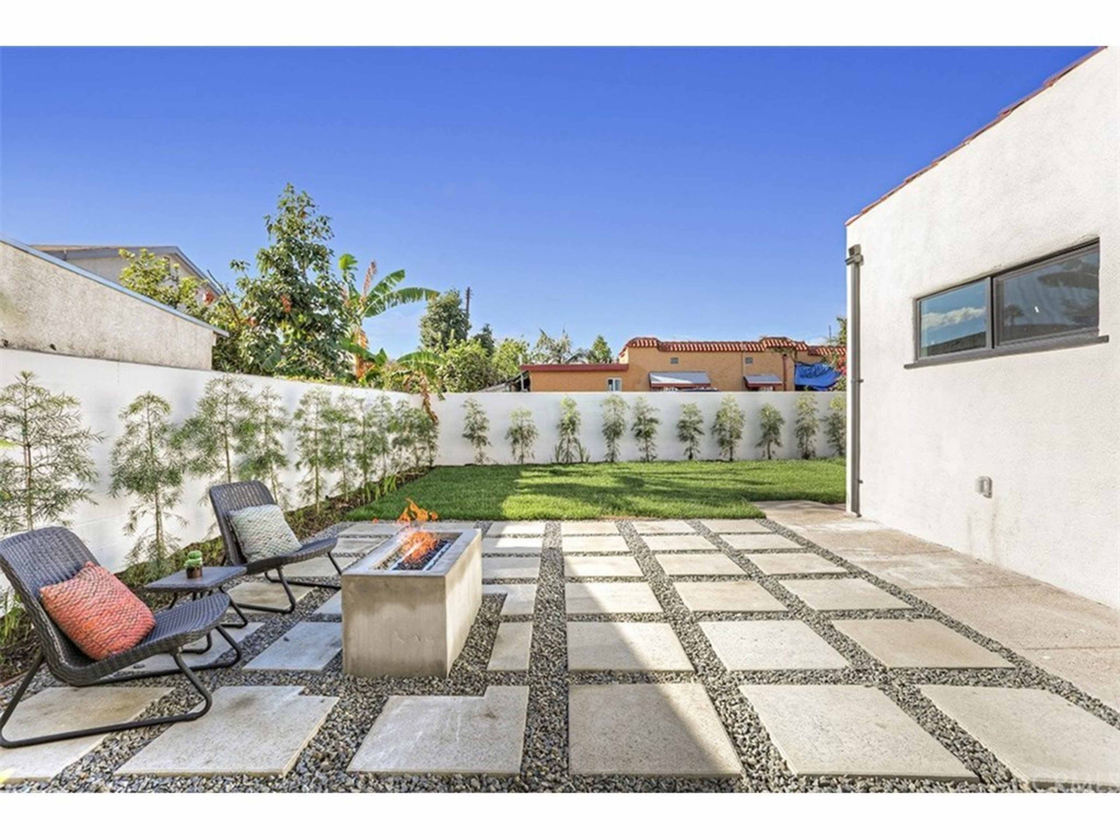 The image depicts a backyard with a stone patio, a fire pit surrounded by chairs, and a green lawn enclosed by white walls and vegetation.