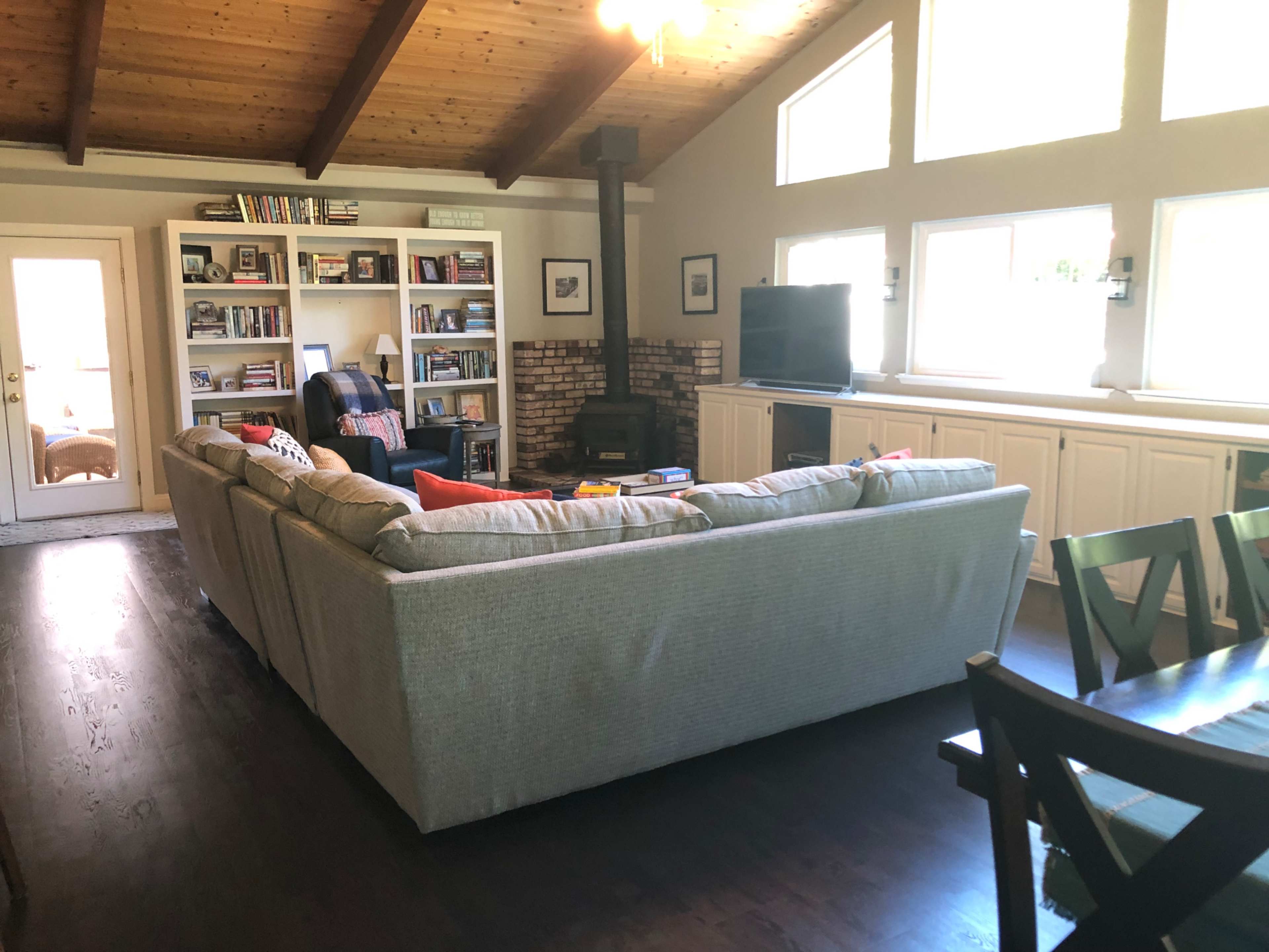 A living room with a large sectional sofa, a wooden ceiling, bookshelves filled with books, and a television next to a wood stove.