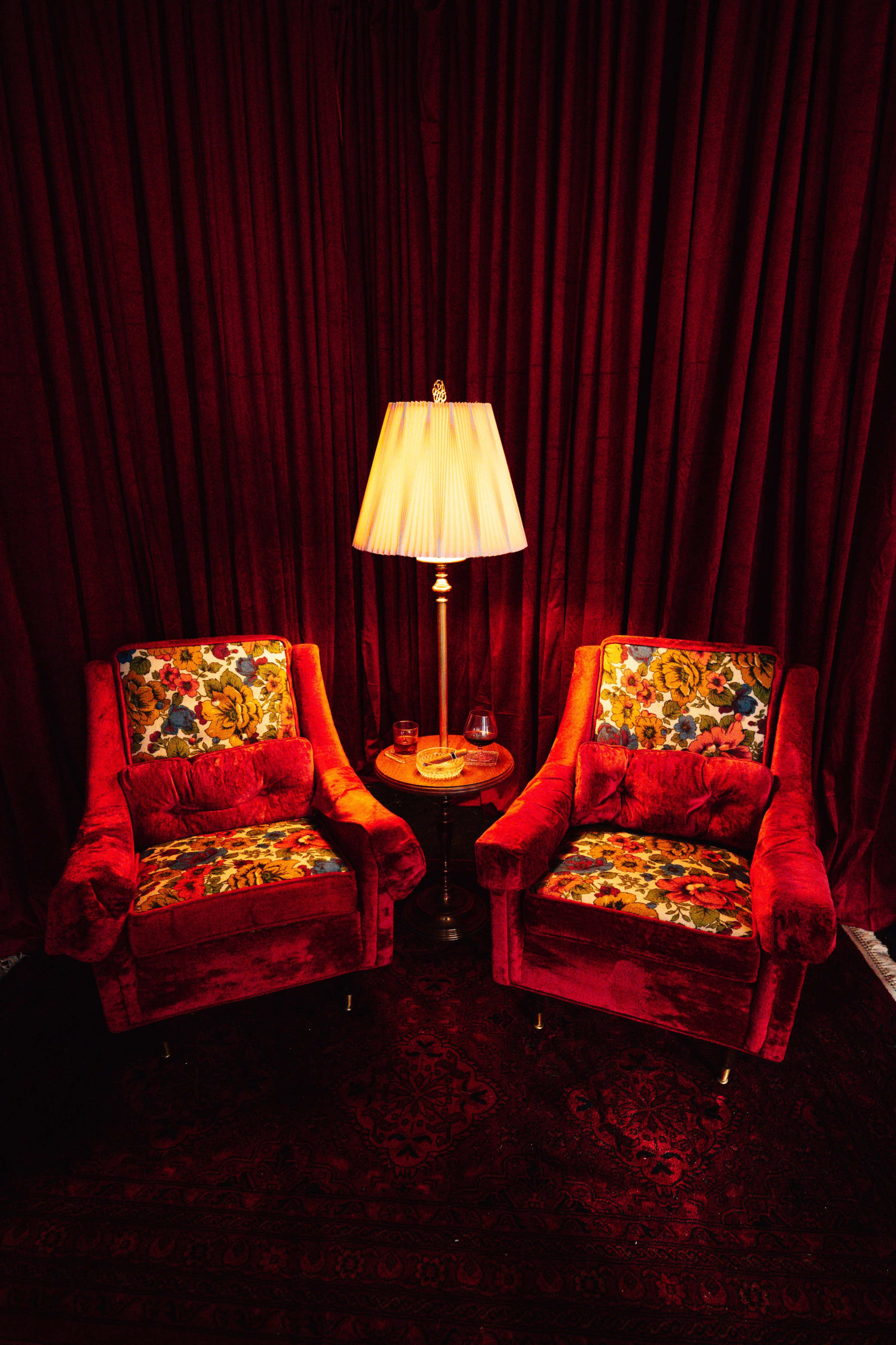 Two velvet armchairs with floral upholstery are positioned beside a small table, topped by a lamp, in front of a deep red curtain.